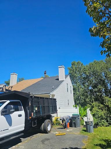 Worker on ladder beside a house roof with a dump truck and tools in the driveway