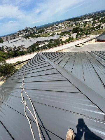 Rooftop view with gray metal roof panels, a person’s shoe, and a cityscape in the distance under a blue sky