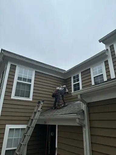 Worker on a roof using a ladder beside a beige house.