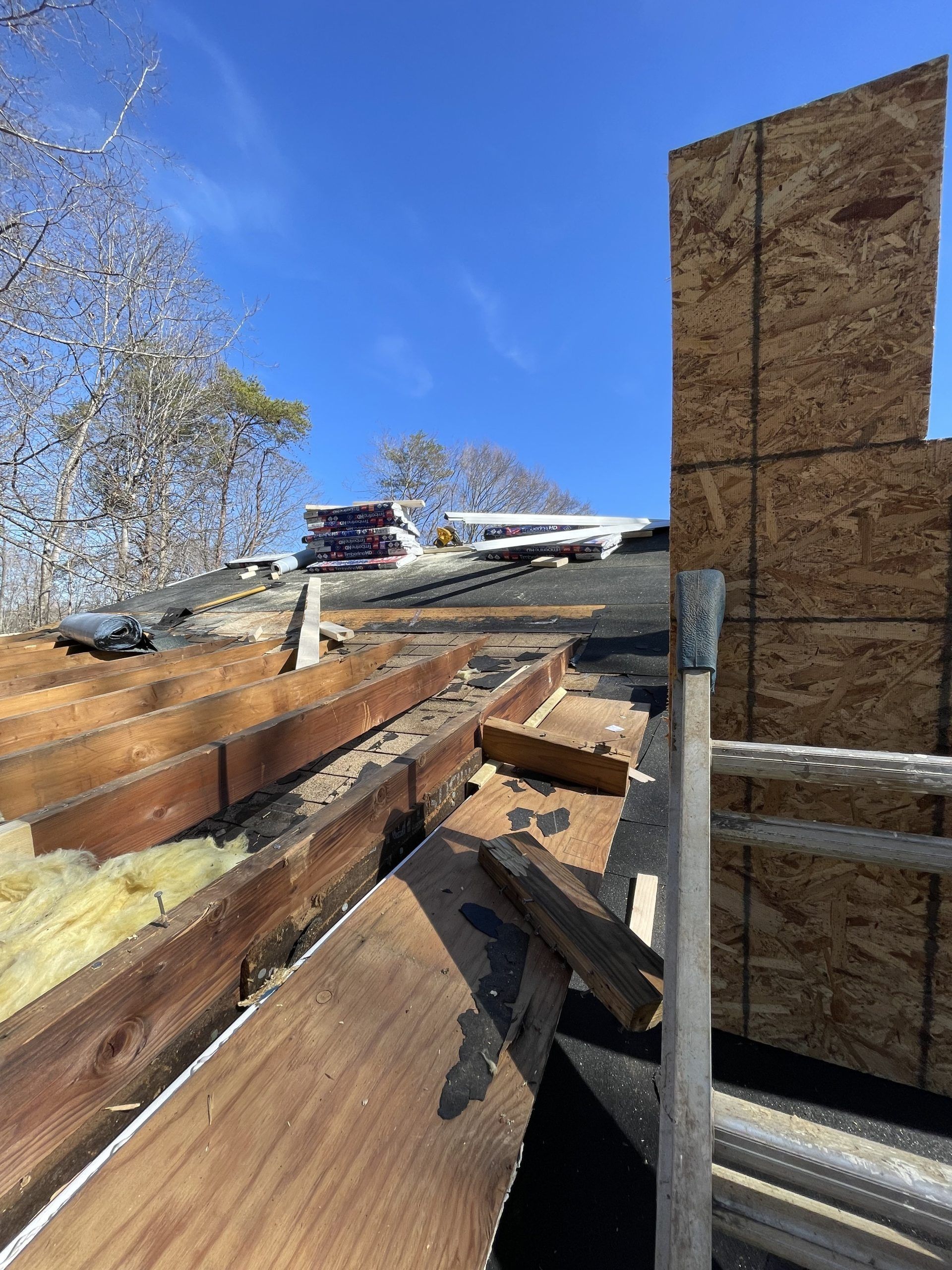 Damaged roof with exposed wooden beams under a blue sky beside a plywood wall
