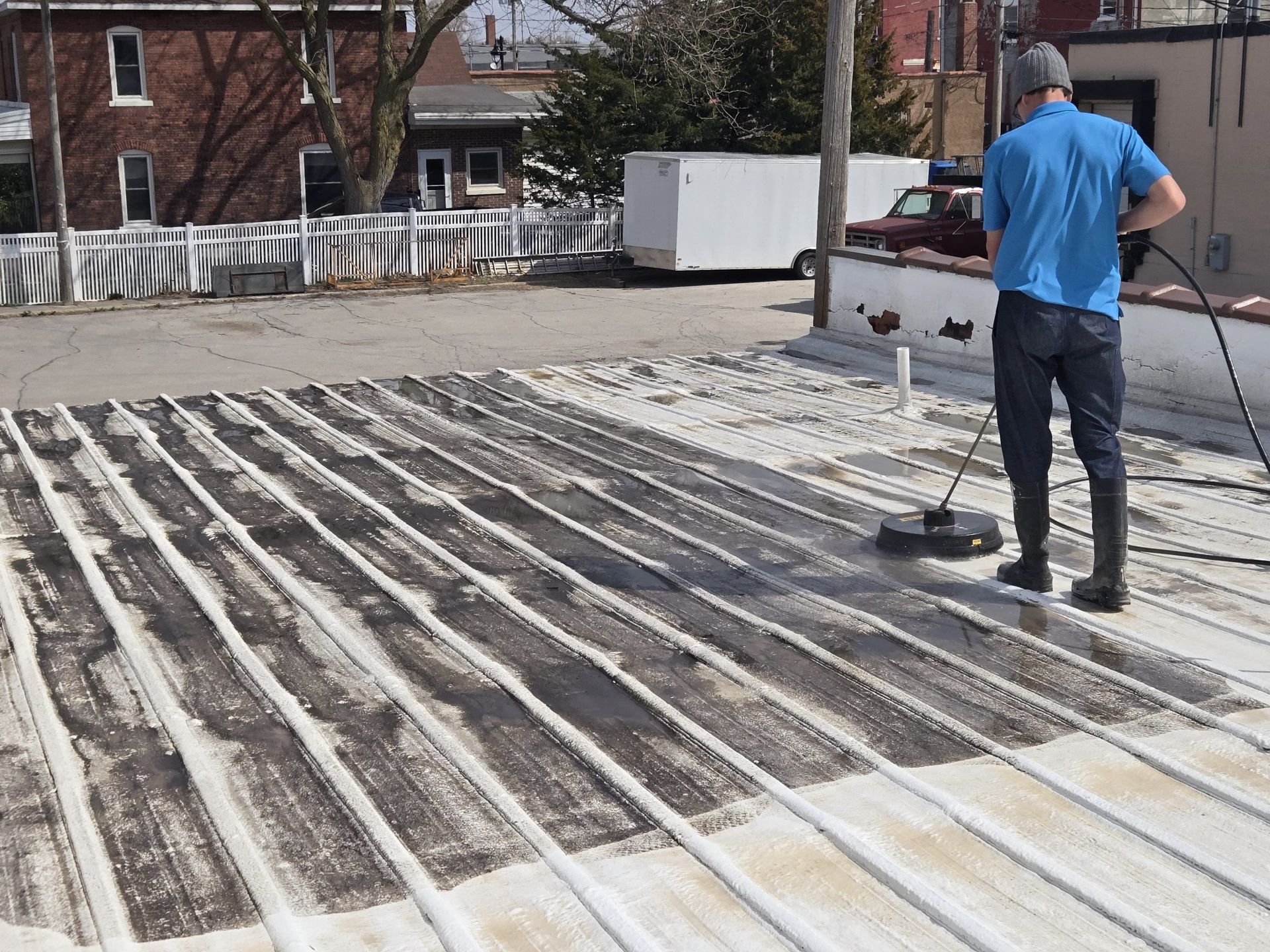 Worker in blue shirt smoothing a rooftop surface with long strips and a tool near a white fence.