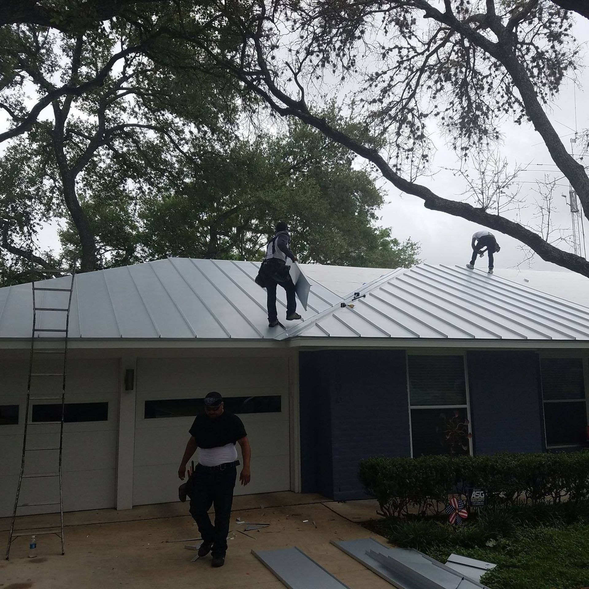 Three workers on a metal roof installing sheets above a house with trees overhead.