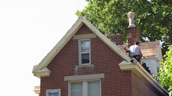 Brick house roof being repaired by a worker near a chimney, with green trees behind.