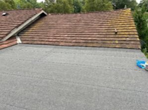 Flat roof under construction beside a tiled roof, with gray roofing membrane and blue tools nearby