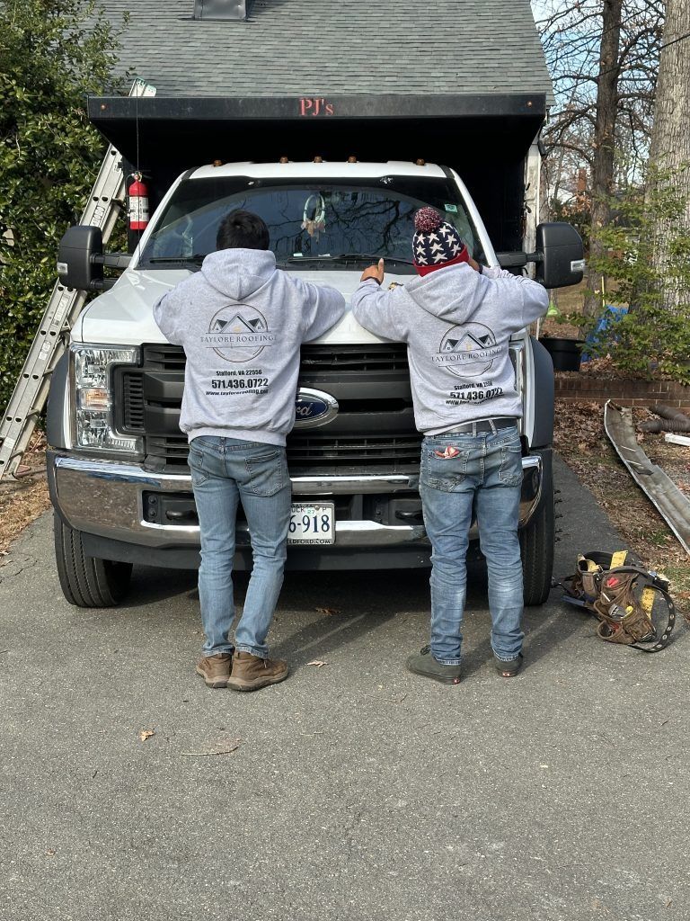 Two people in hoodies stand at the front of a pickup truck in a driveway, washing its windshield.