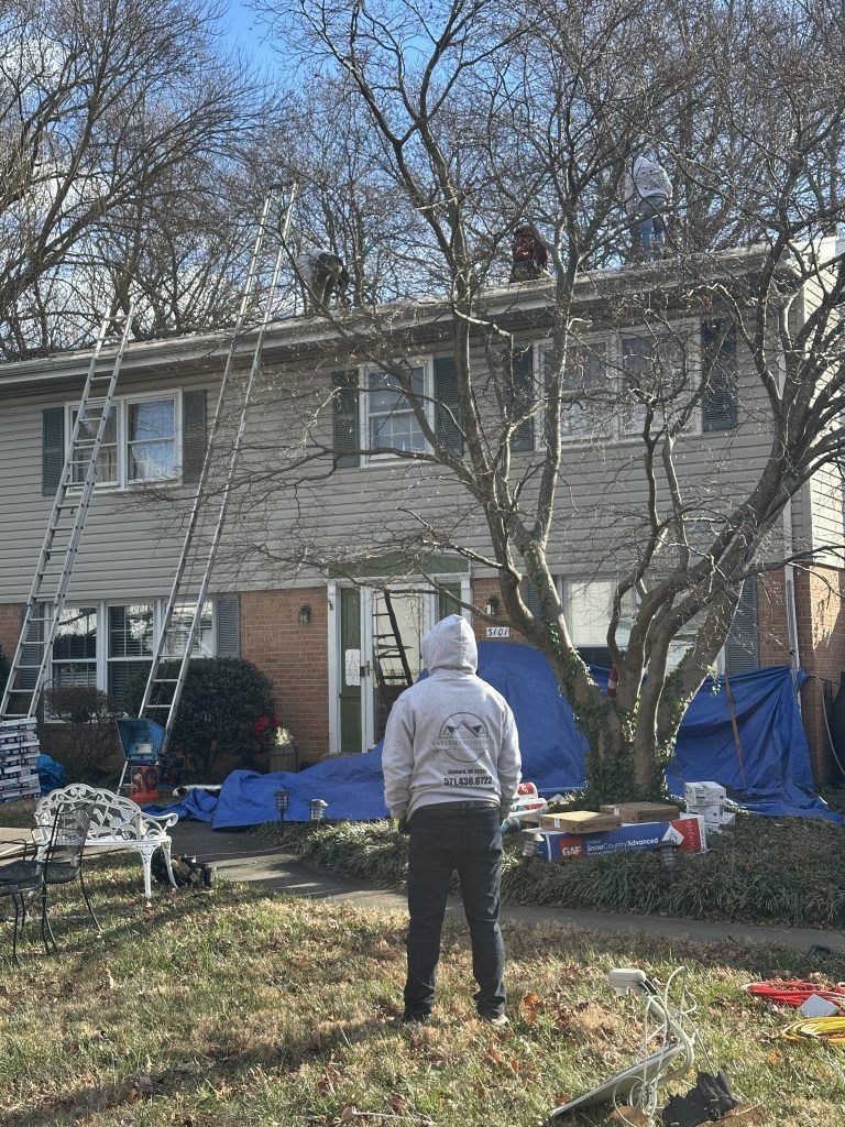 Person stands in a yard near a two-story house with ladders and blue tarps during exterior work.