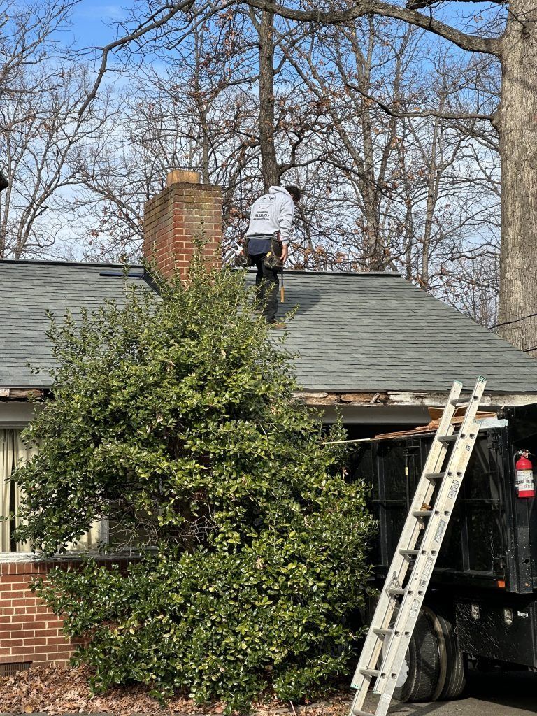Worker on a house roof near a chimney, with a ladder and tree in front.