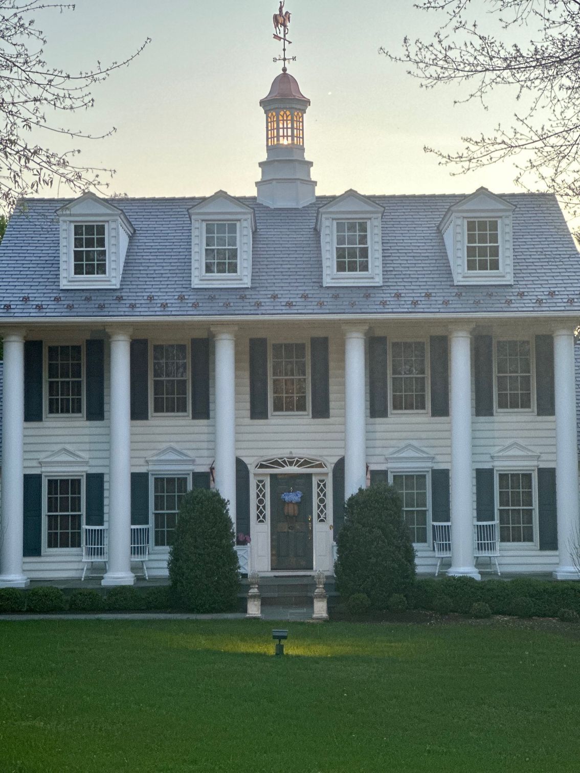 Large white colonial-style house with columns, dormer windows, and a central cupola behind a green lawn.
