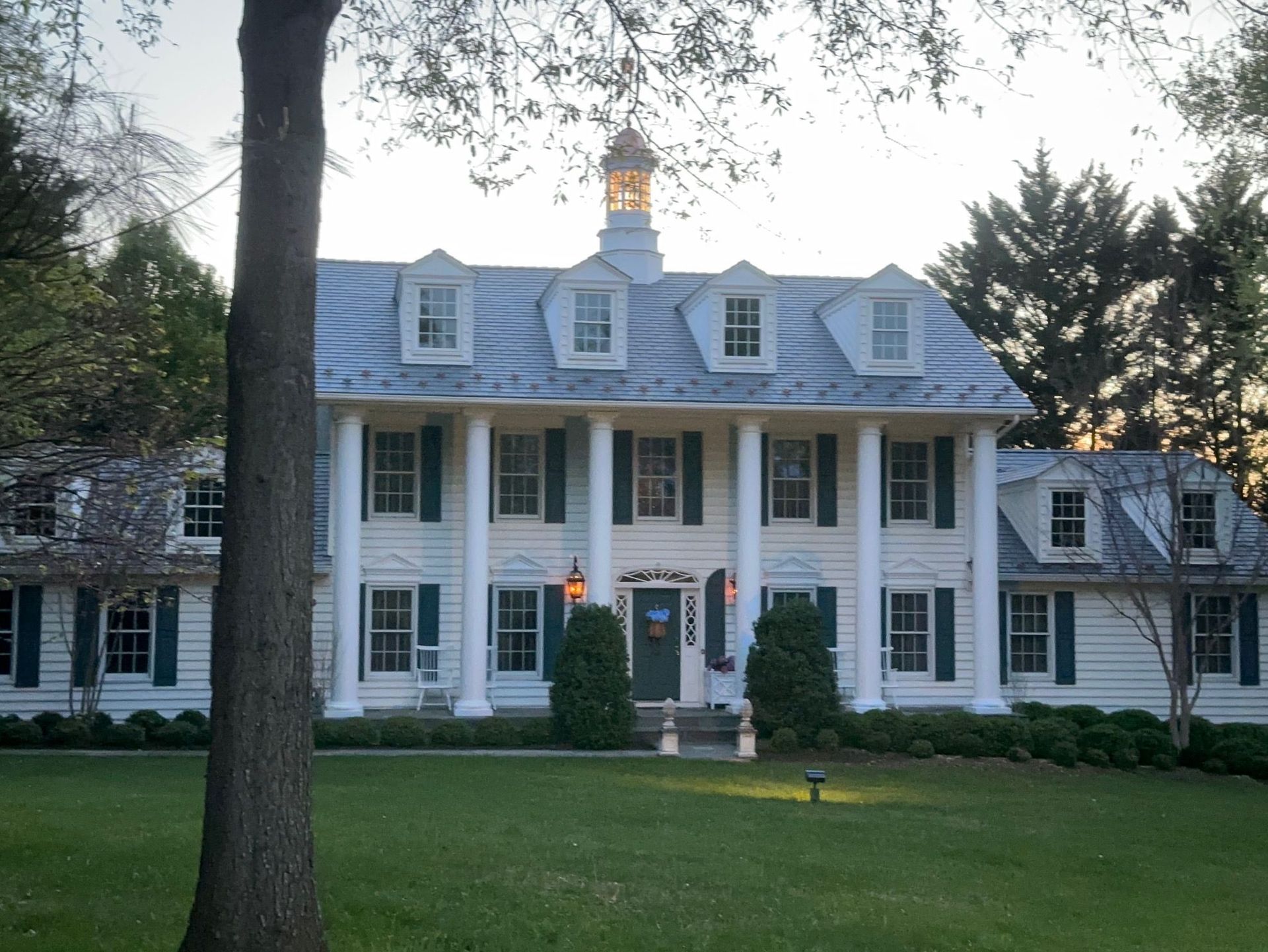 Large white colonial mansion with columns, lit porch, and manicured lawn at dusk