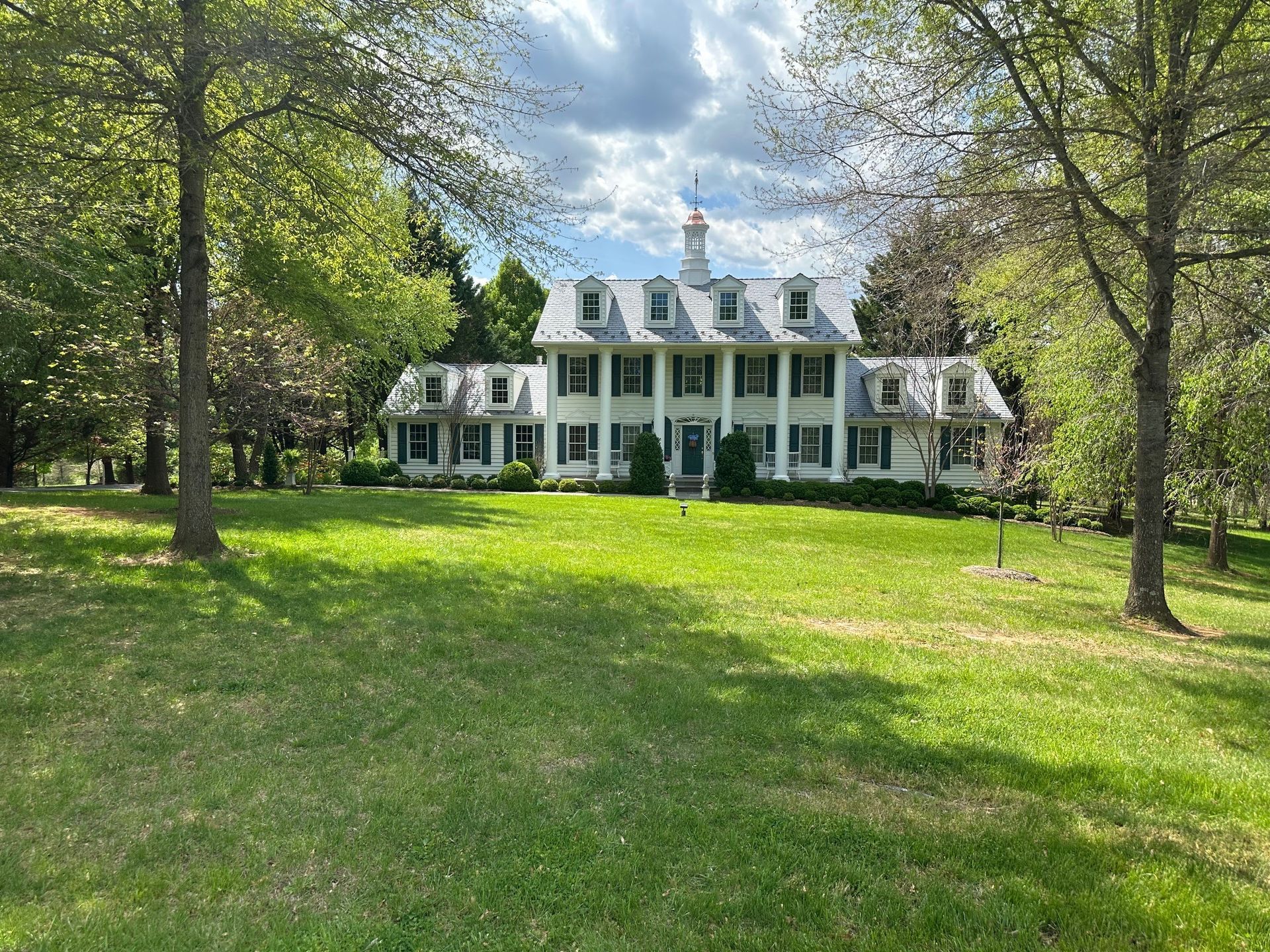 White colonial-style house with green shutters on a sunny lawn, framed by trees