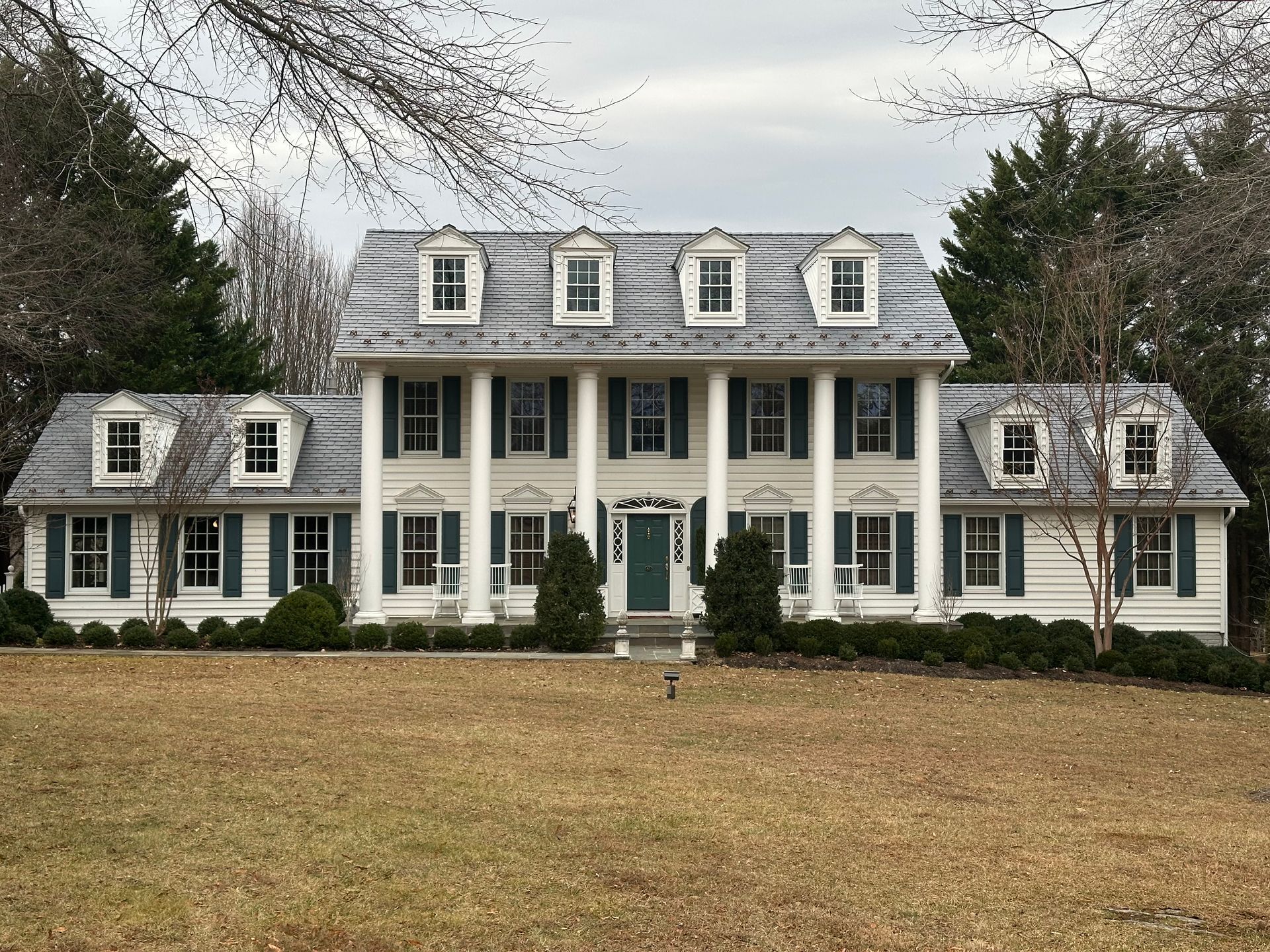 Large white colonial-style house with tall columns, dormer windows, and a wide lawn under cloudy skies
