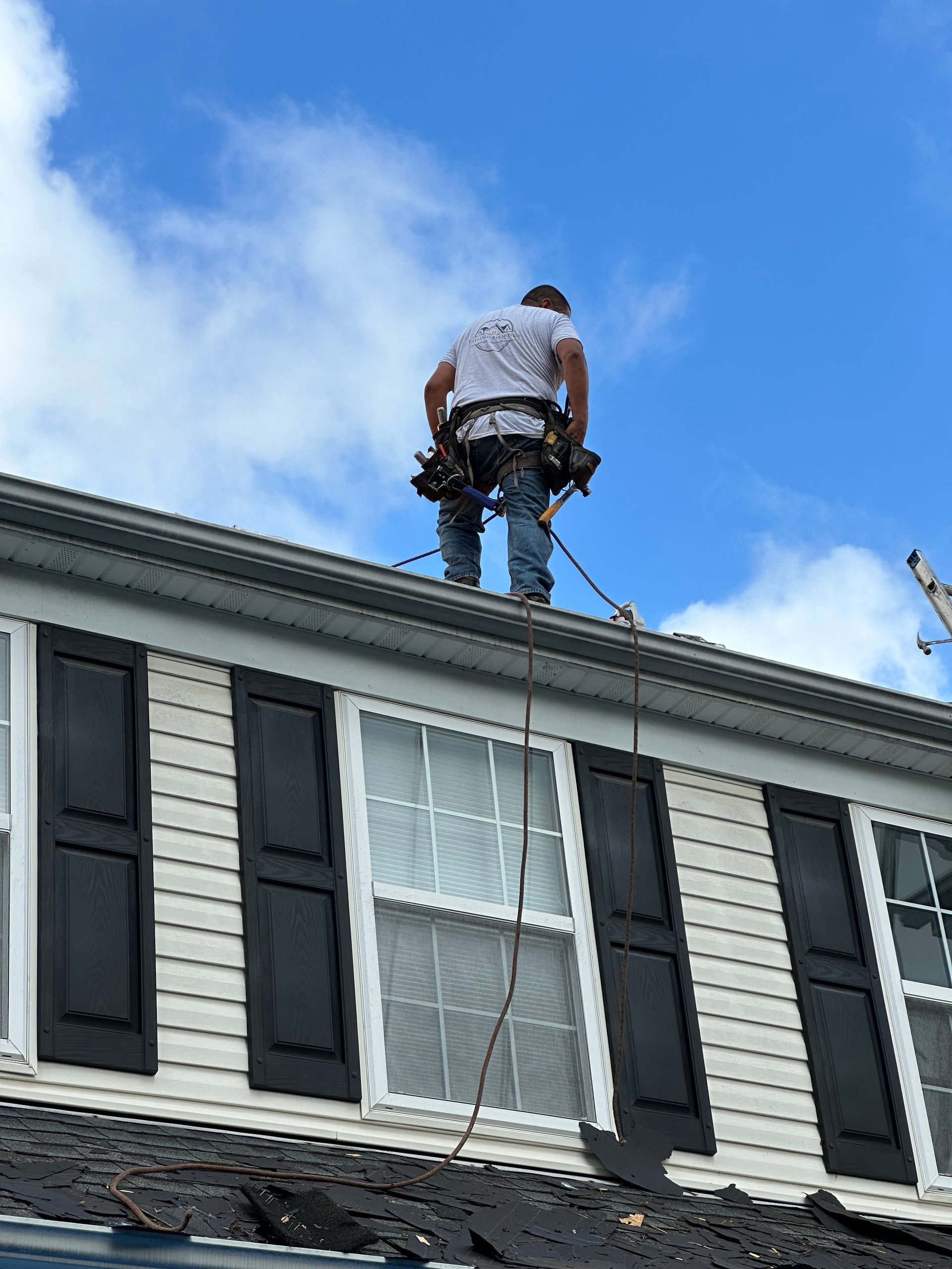 Worker on a roof beside black shutters, securing equipment against a blue sky.