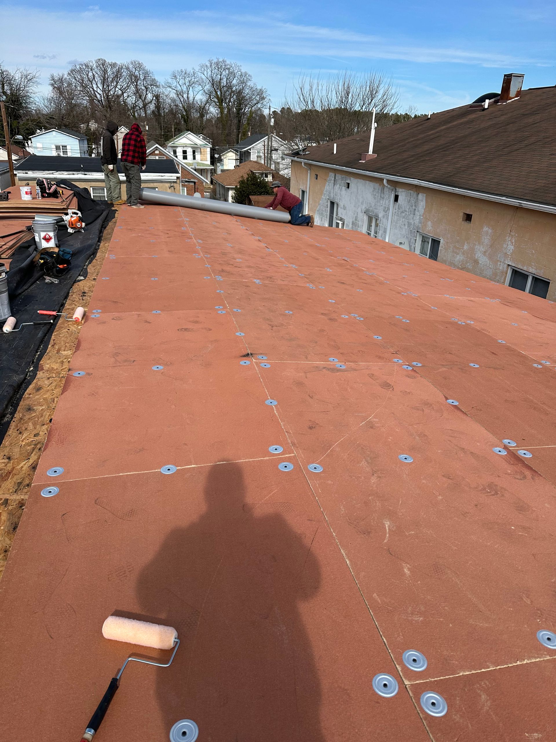 Workers on a flat roof applying orange roofing material under a clear blue sky.
