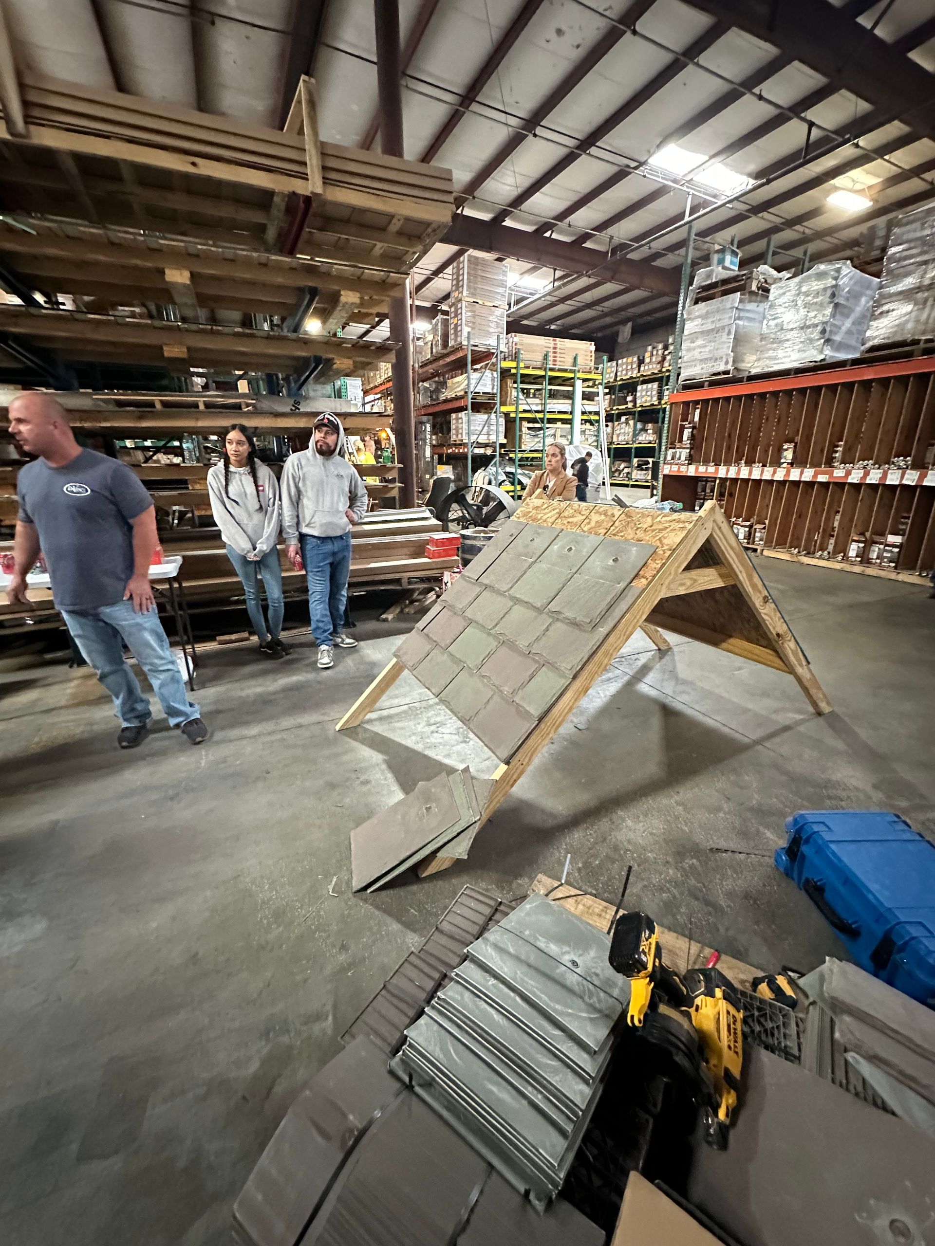 People in a warehouse workshop beside a ramp-like structure and stacked tools and materials.