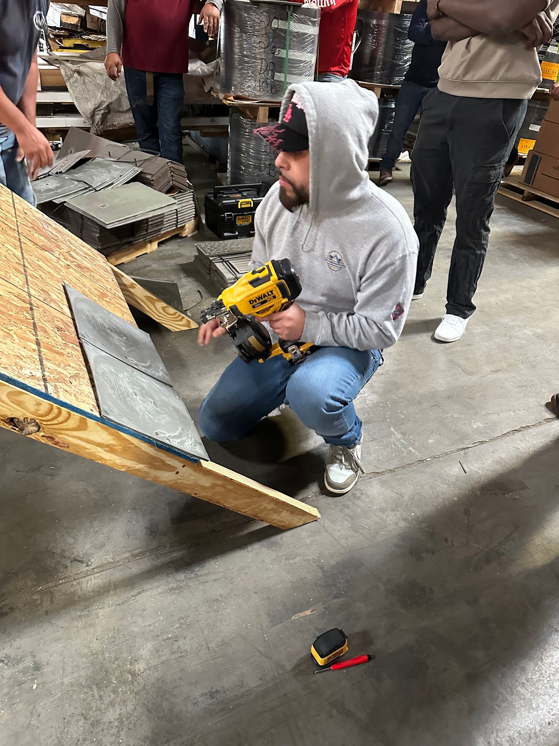 Person in gray hoodie crouches on a wet street, holding a yellow tool while examining a tilted wooden panel.