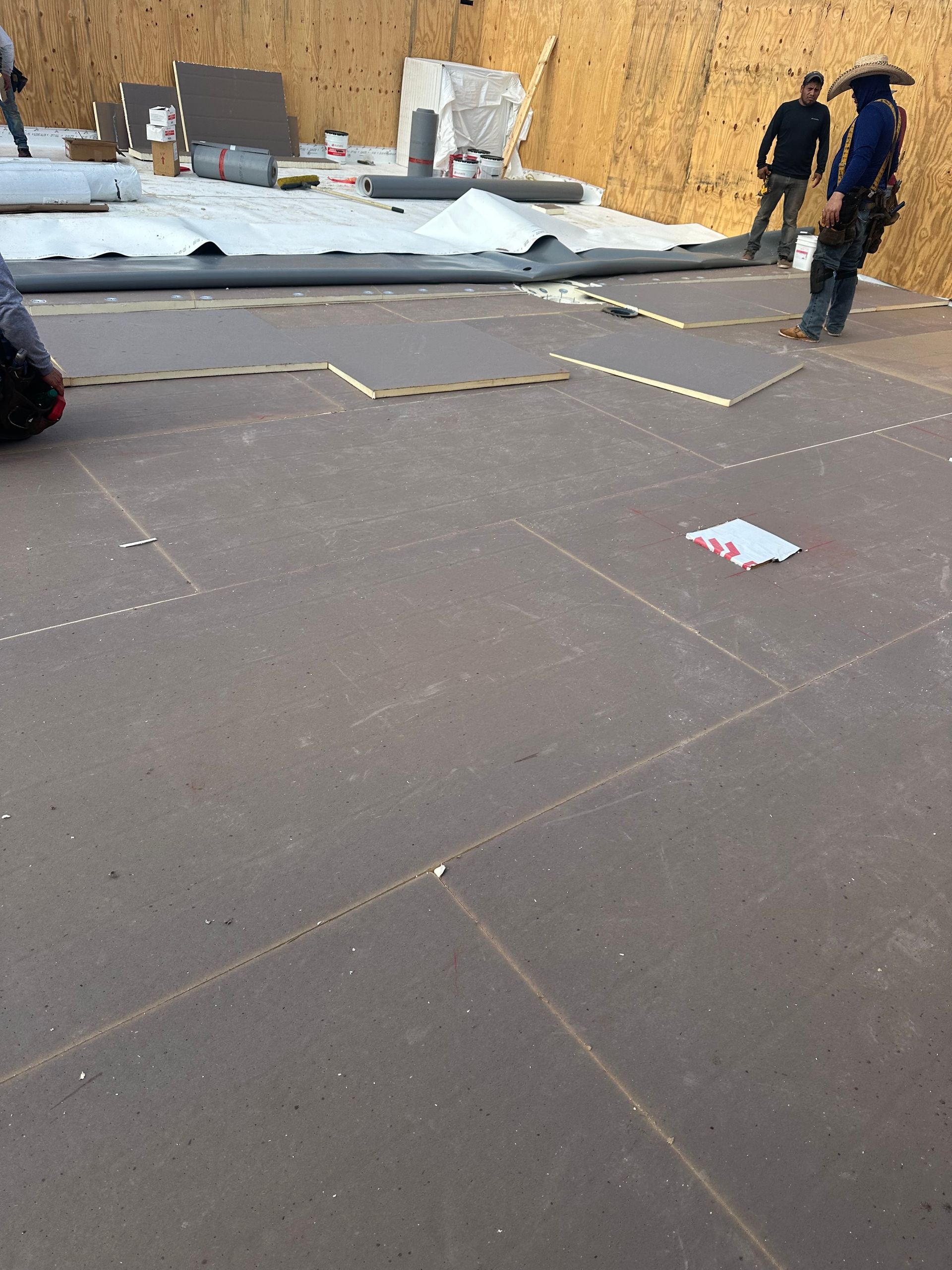 Roofing workers laying materials on a flat roof, with white insulation sheets and plywood in the background.