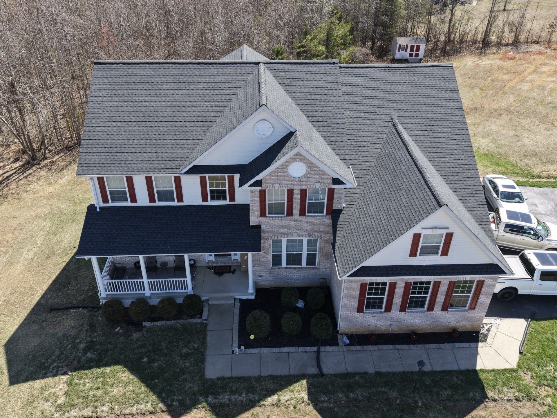 Aerial view of a large white house with a dark roof, porch, and driveway in a wooded yard.