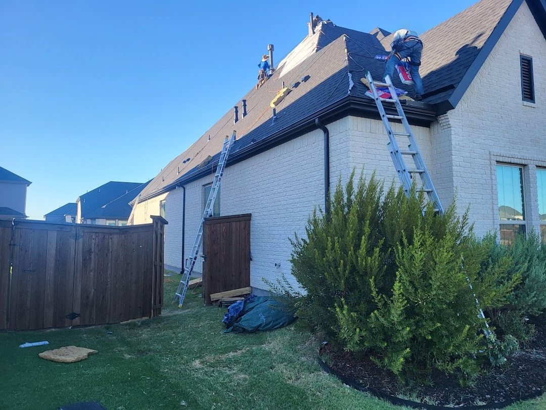 House exterior with workers on the roof and a ladder leaned against the wall, beside a fence and shrubs