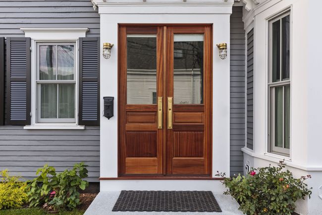 Wooden double front doors with glass panels on a gray house, framed by white trim and black wall lanterns