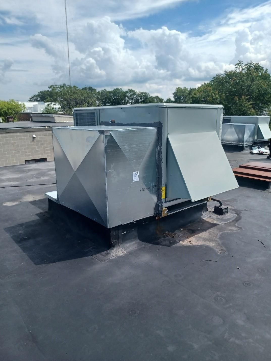HVAC rooftop unit on a flat, black surface against a partly cloudy blue sky.