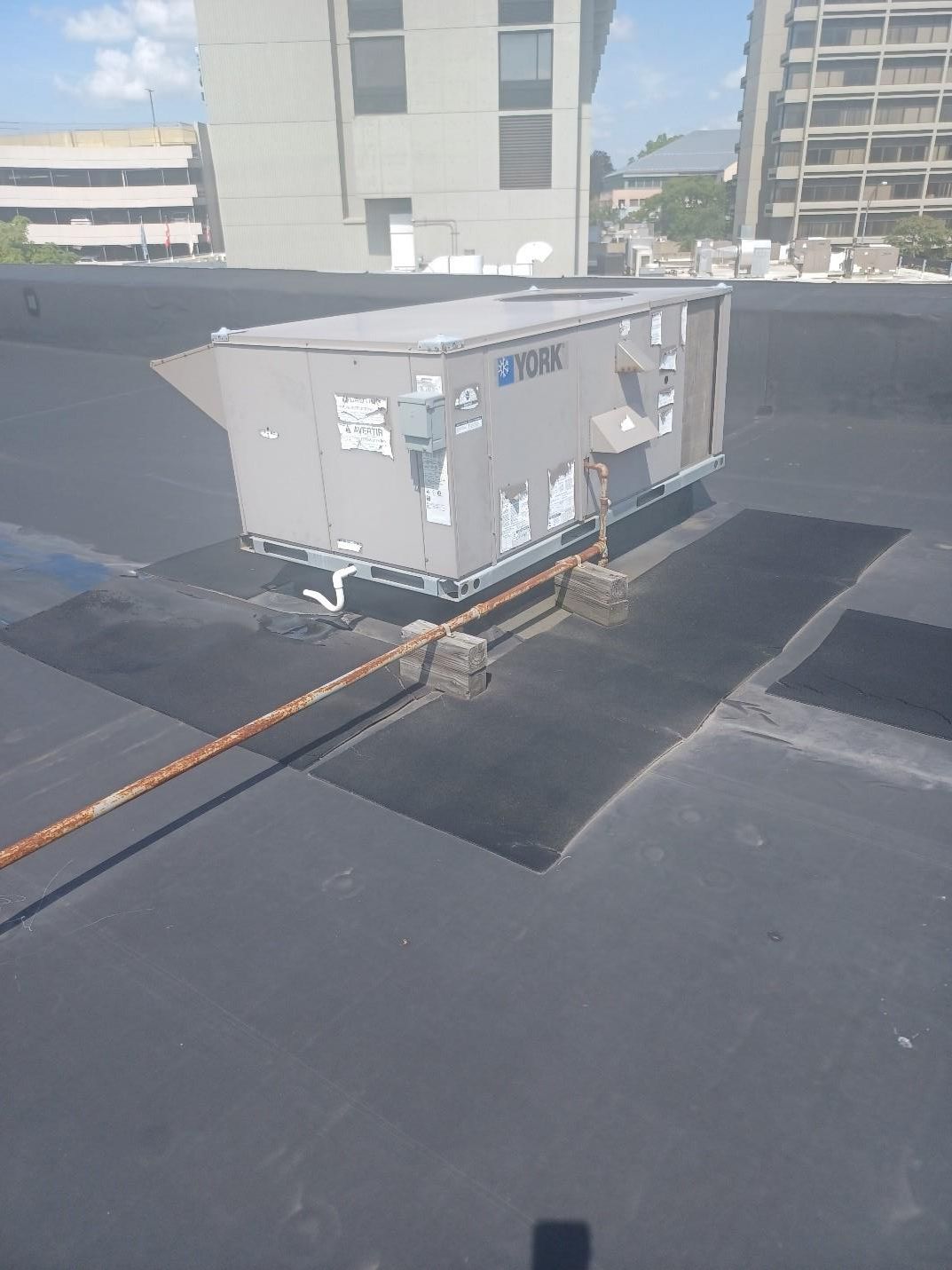 HVAC unit on a flat, black rooftop, with metal supports and a rusty cable running along the surface.