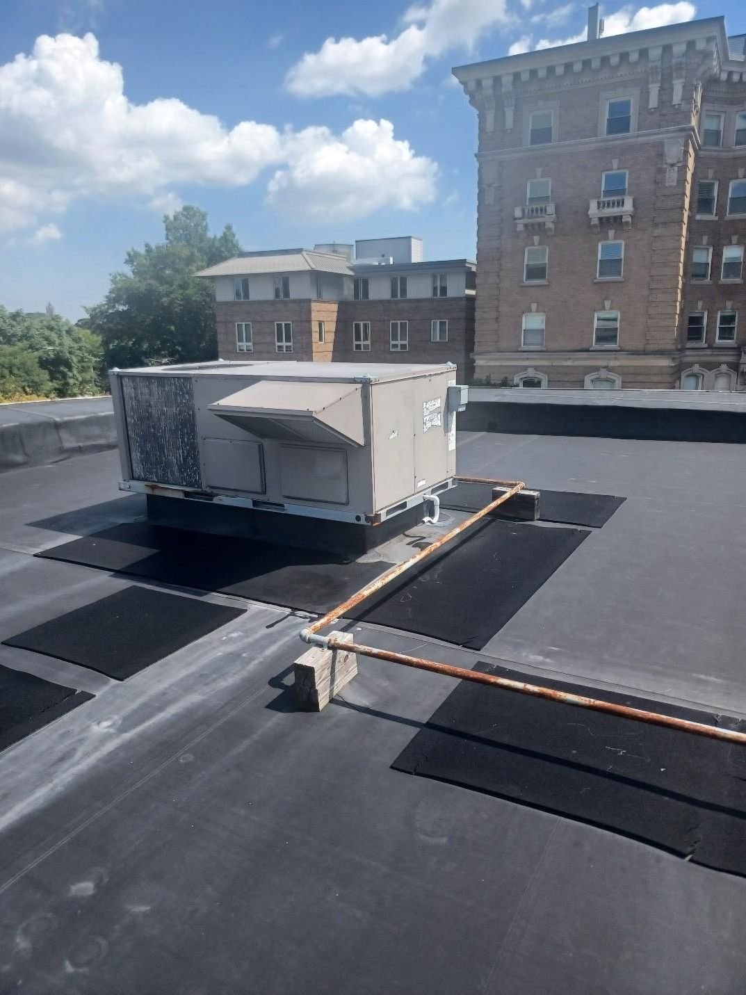 Rooftop with HVAC unit, copper pipes, and adjacent brick building under a cloudy sky.