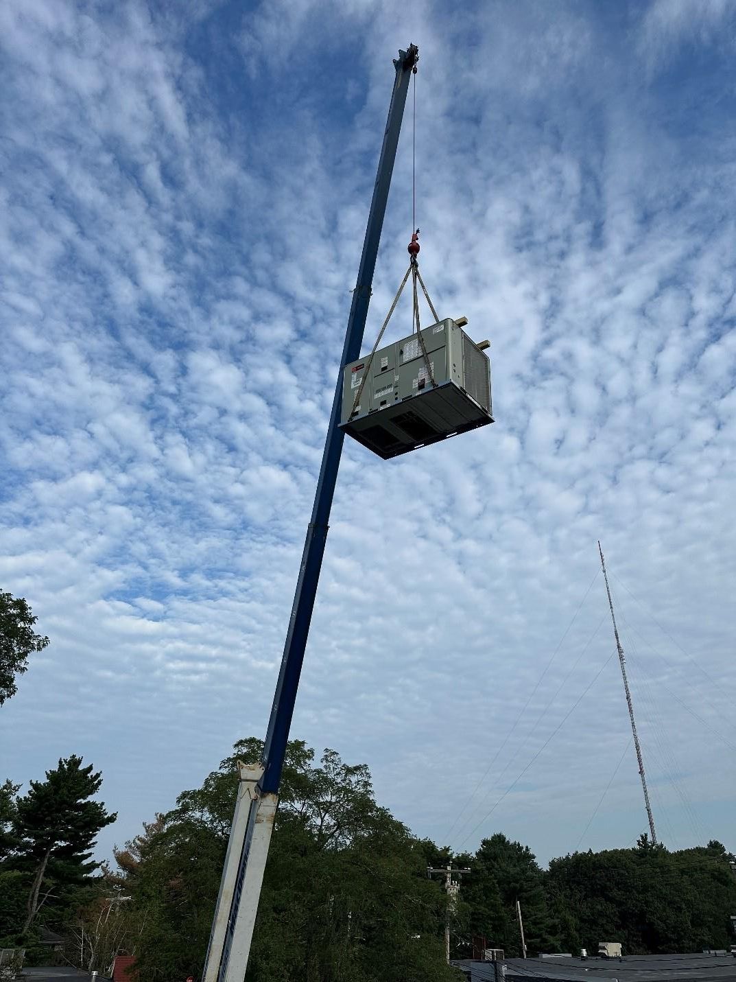 A crane lifts a large rectangular HVAC unit against a partly cloudy sky.