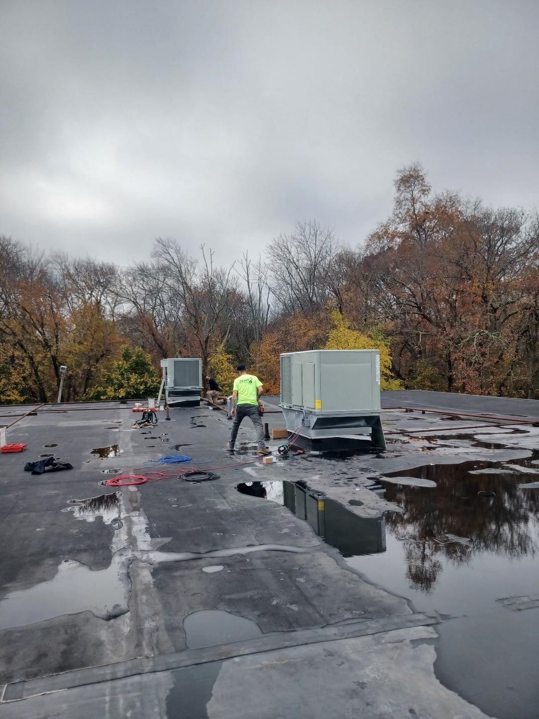A person in a yellow vest works on rooftop AC units on a wet, cloudy day.