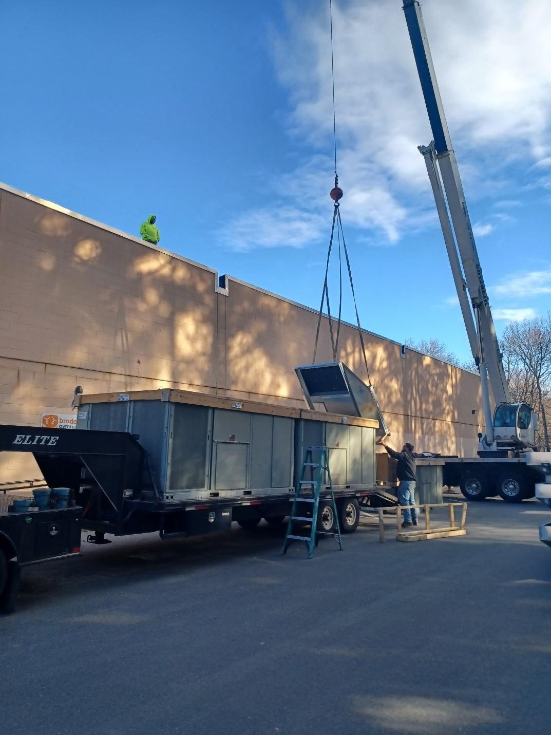 A crane lifting a large, rectangular HVAC unit from a flatbed truck onto a building's roof.