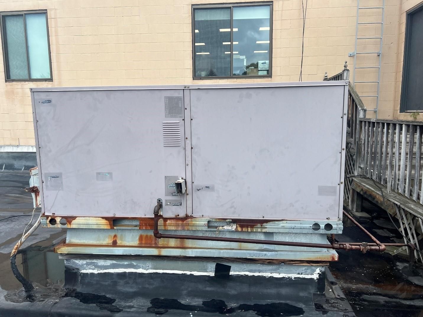 Rooftop air conditioning unit, gray and rusty, against a light brick building and sky.
