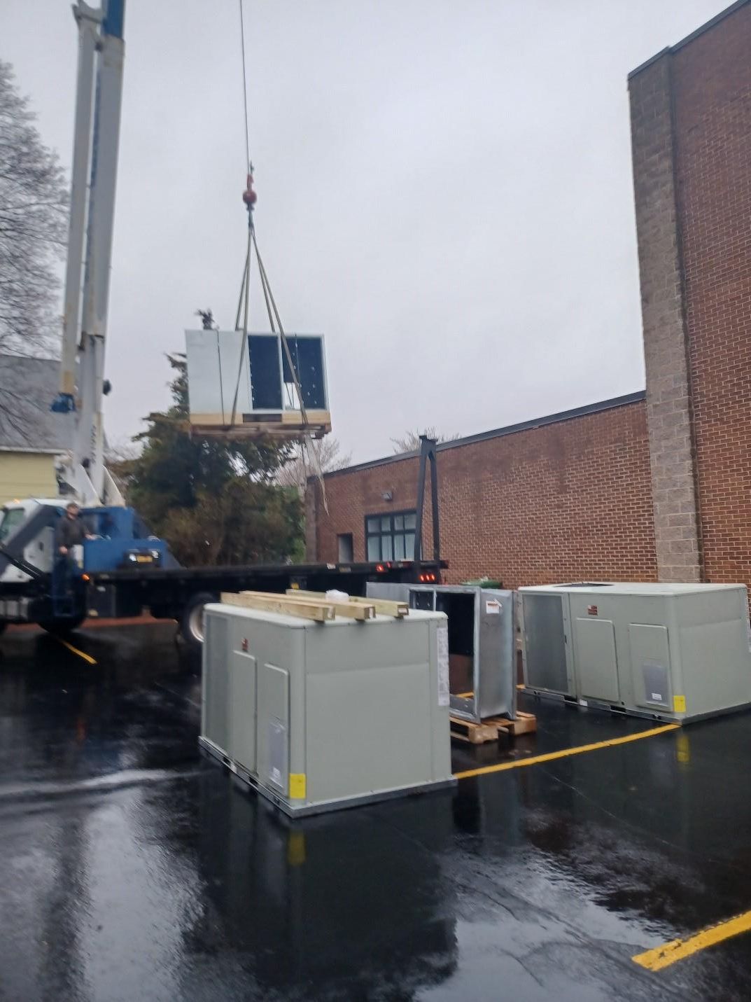 Crane lifting HVAC unit near building, other units on ground in wet conditions.