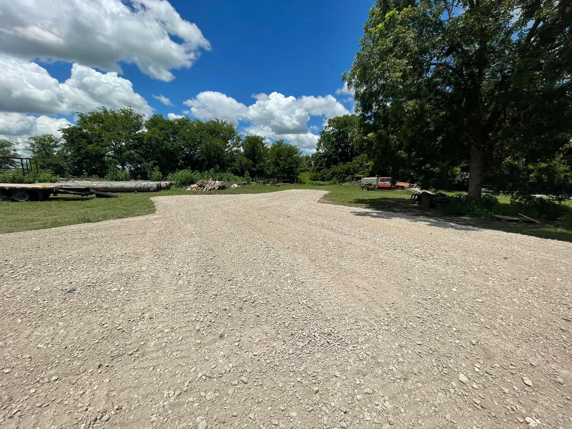 Gravel driveway leading to a grassy area with trees under a blue sky with clouds.
