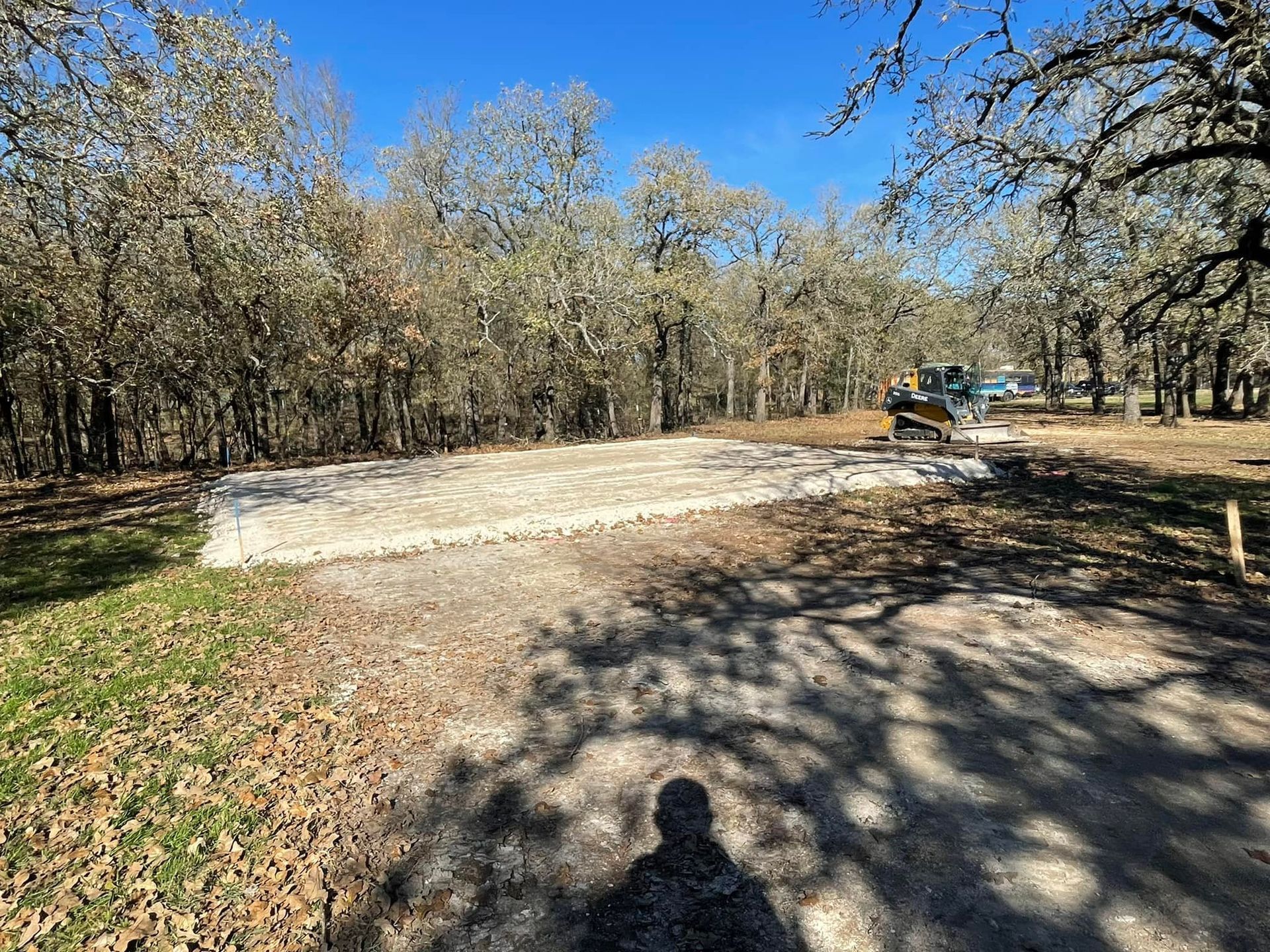 Gravel pad being leveled by machinery in a clearing surrounded by trees, under a blue sky.