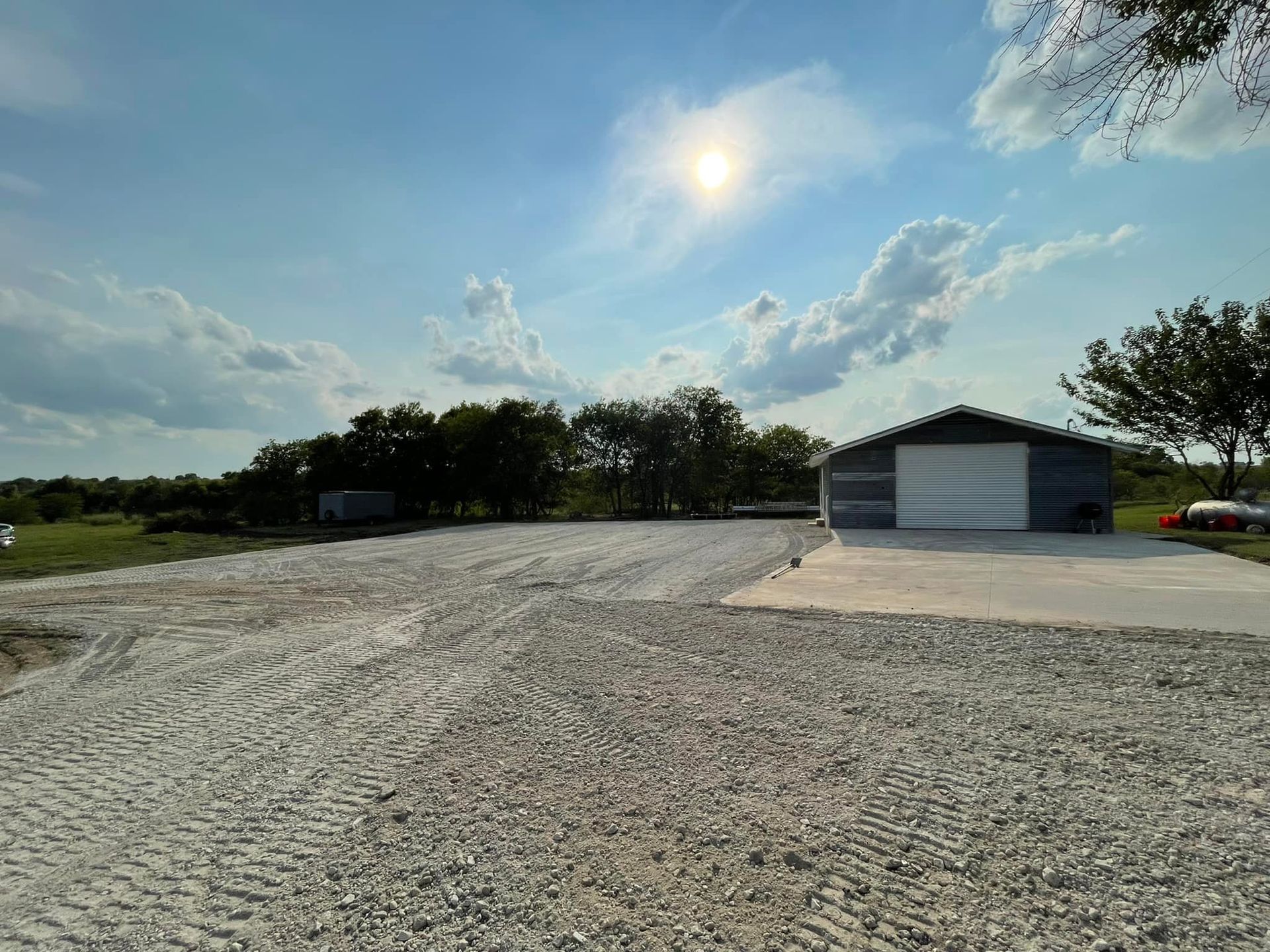 Gravel driveway leading to a gray metal building on a sunny day with trees in the background.
