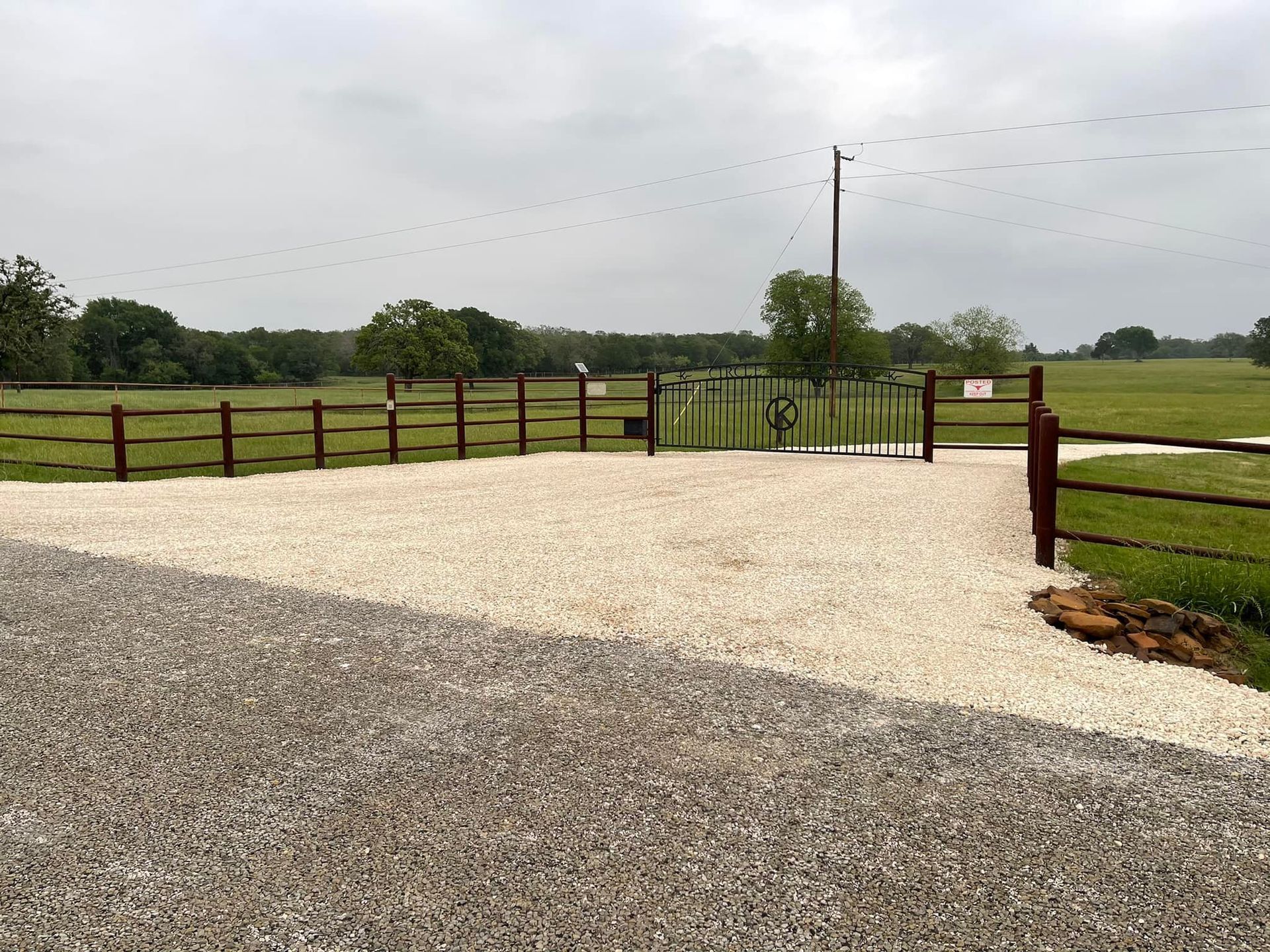 Gravel driveway leads to a fenced property with a gate. Green fields and trees under cloudy sky.