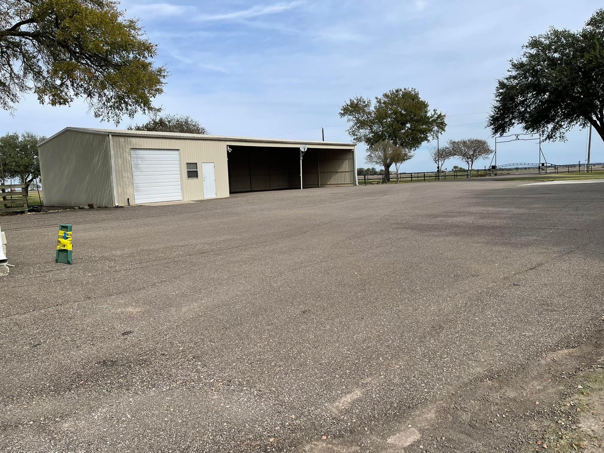 Gravel parking area with a tan, open-sided shed, trees, and a blue sky.