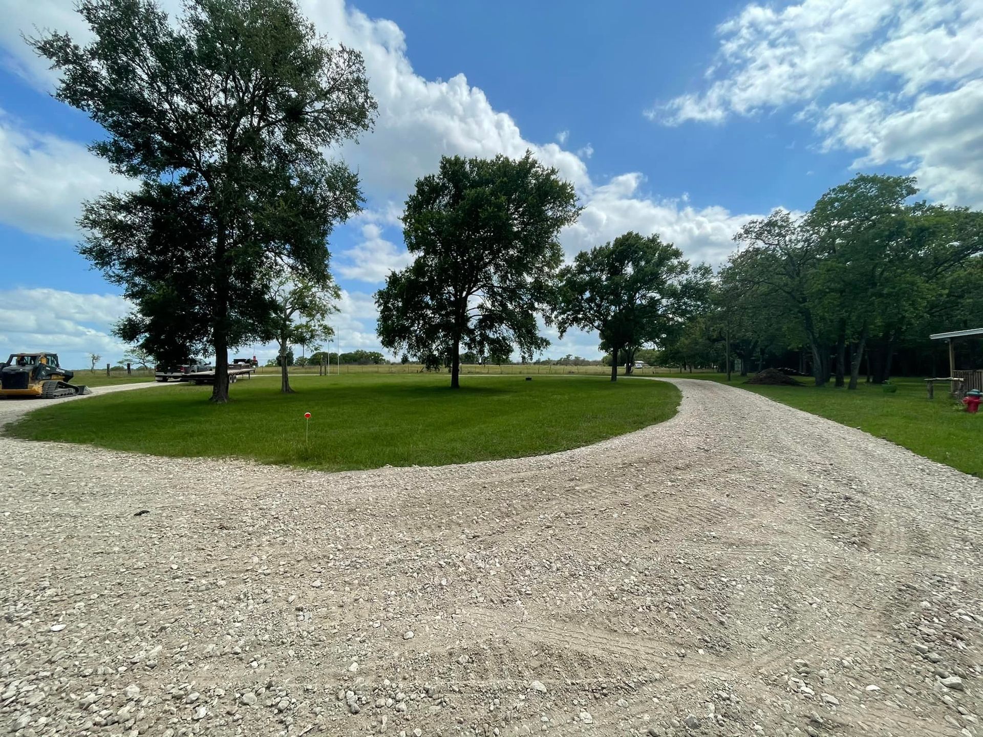Gravel driveway splitting, leading towards green field with scattered trees under a blue sky with clouds.