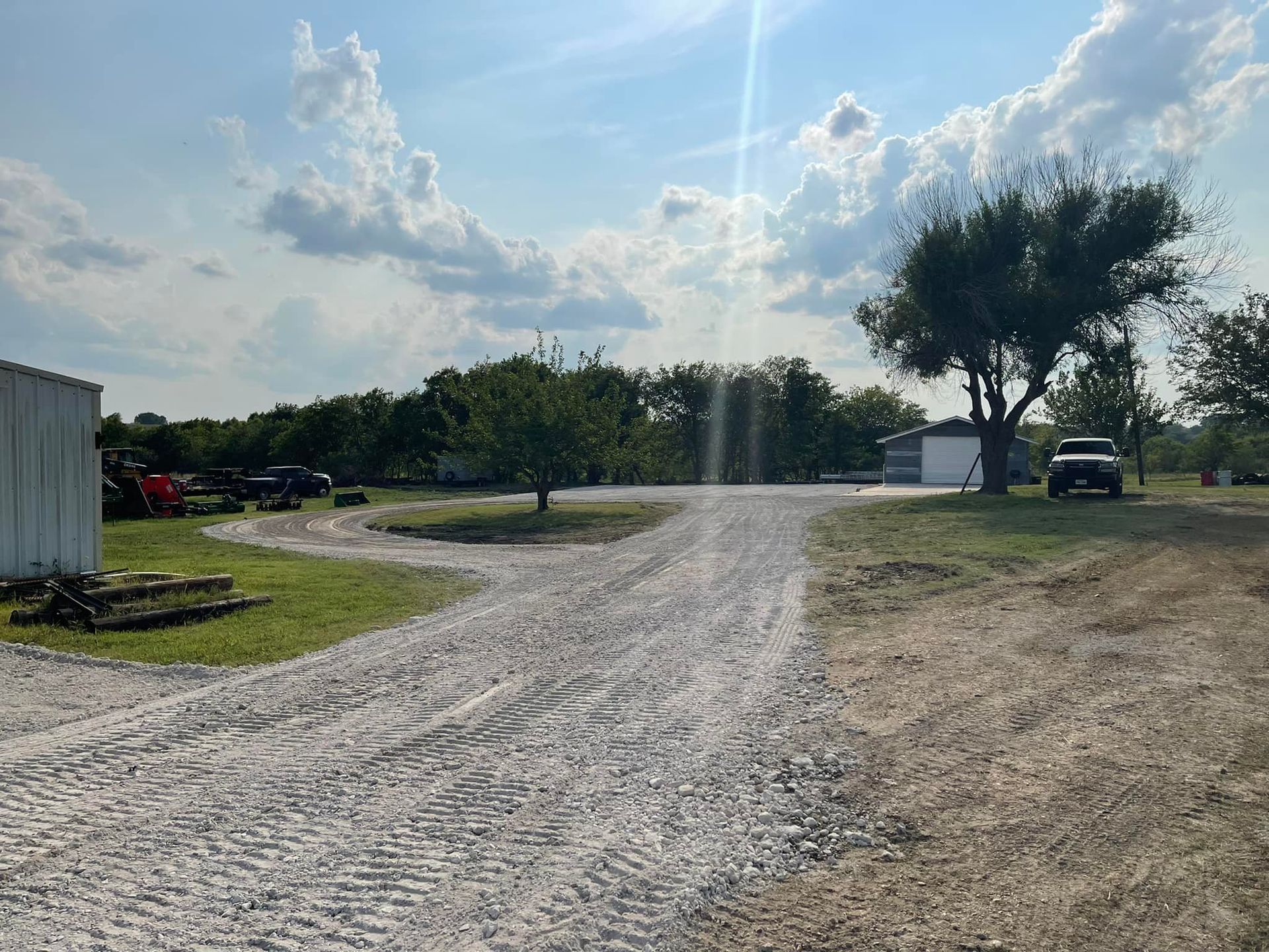 Gravel driveway leading to buildings and trees under a sunny sky.