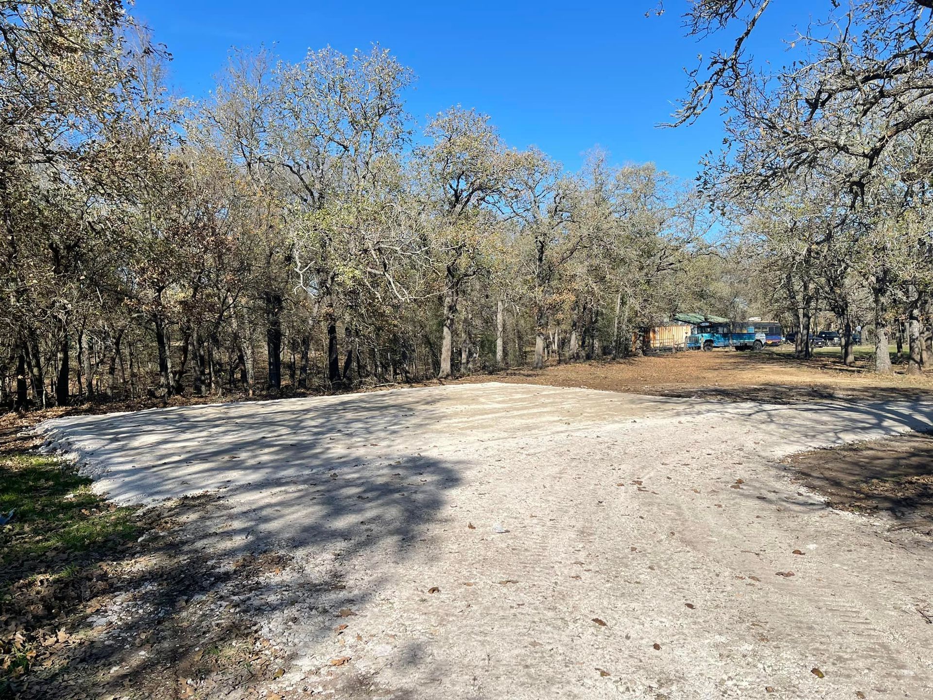 Gravel driveway leading to trees, with a building visible in the distance under a blue sky.