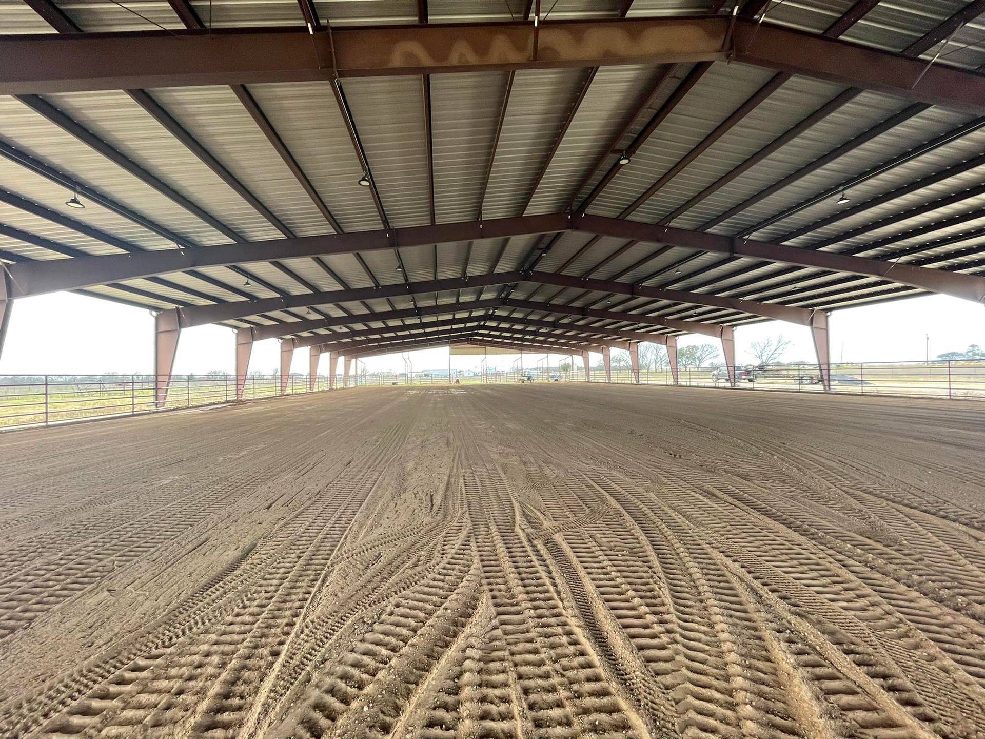 Large, open-air riding arena with tan dirt floor and a metal roof supported by steel beams.