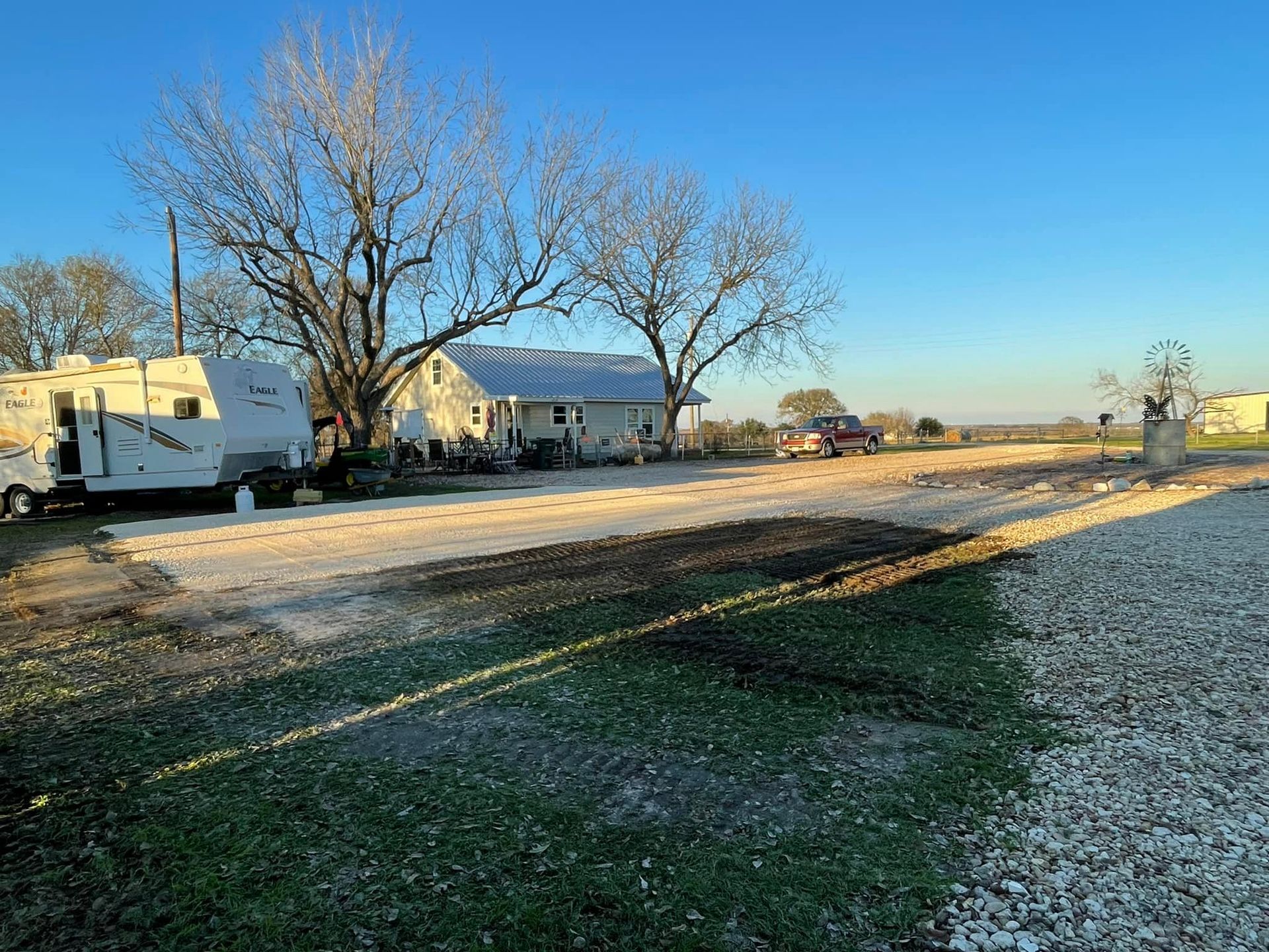RV parked next to a house, gravel road in front, trees and clear sky.