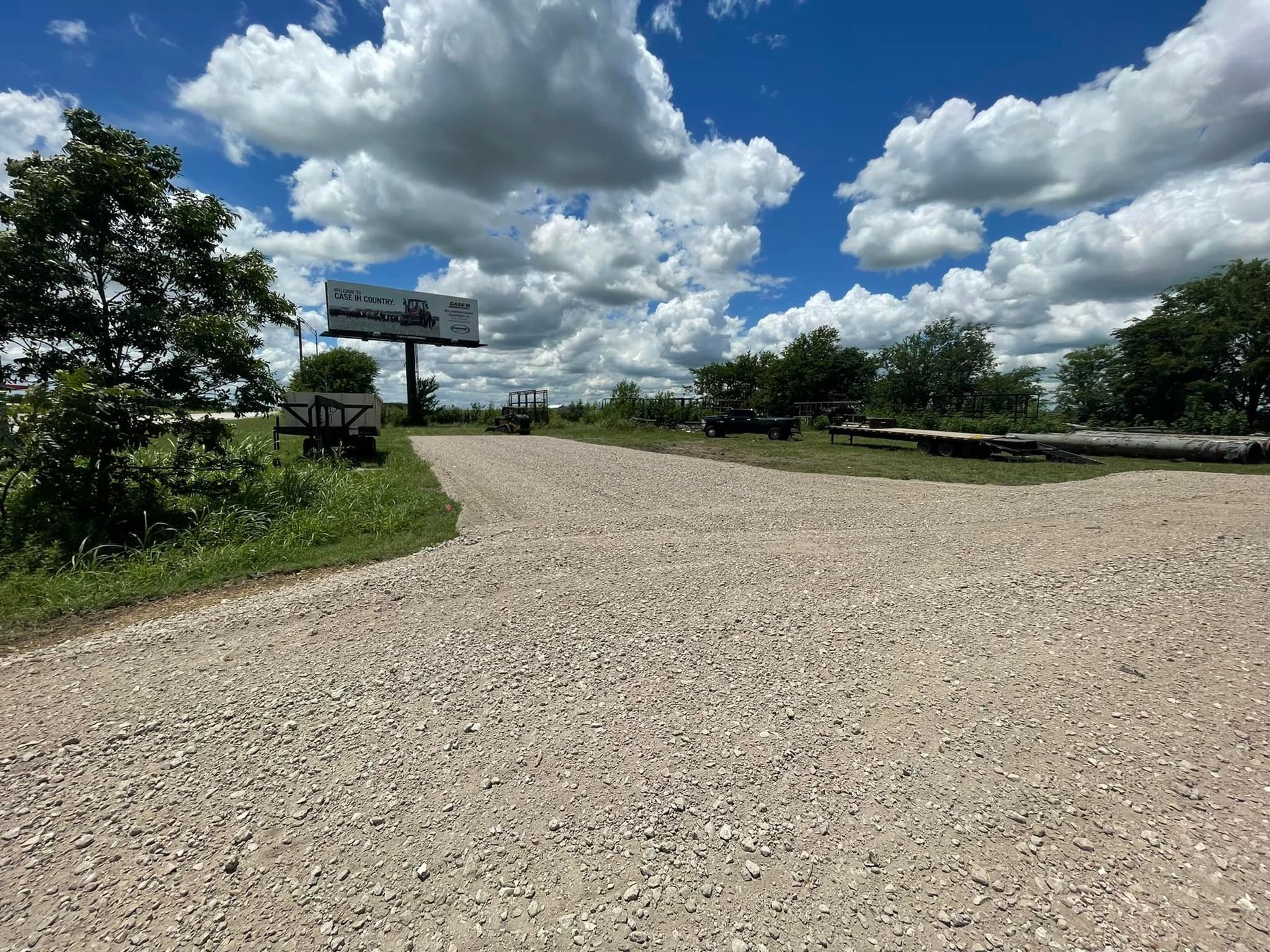 A gravel road leads toward a billboard under a cloudy, blue sky; trees line the sides.