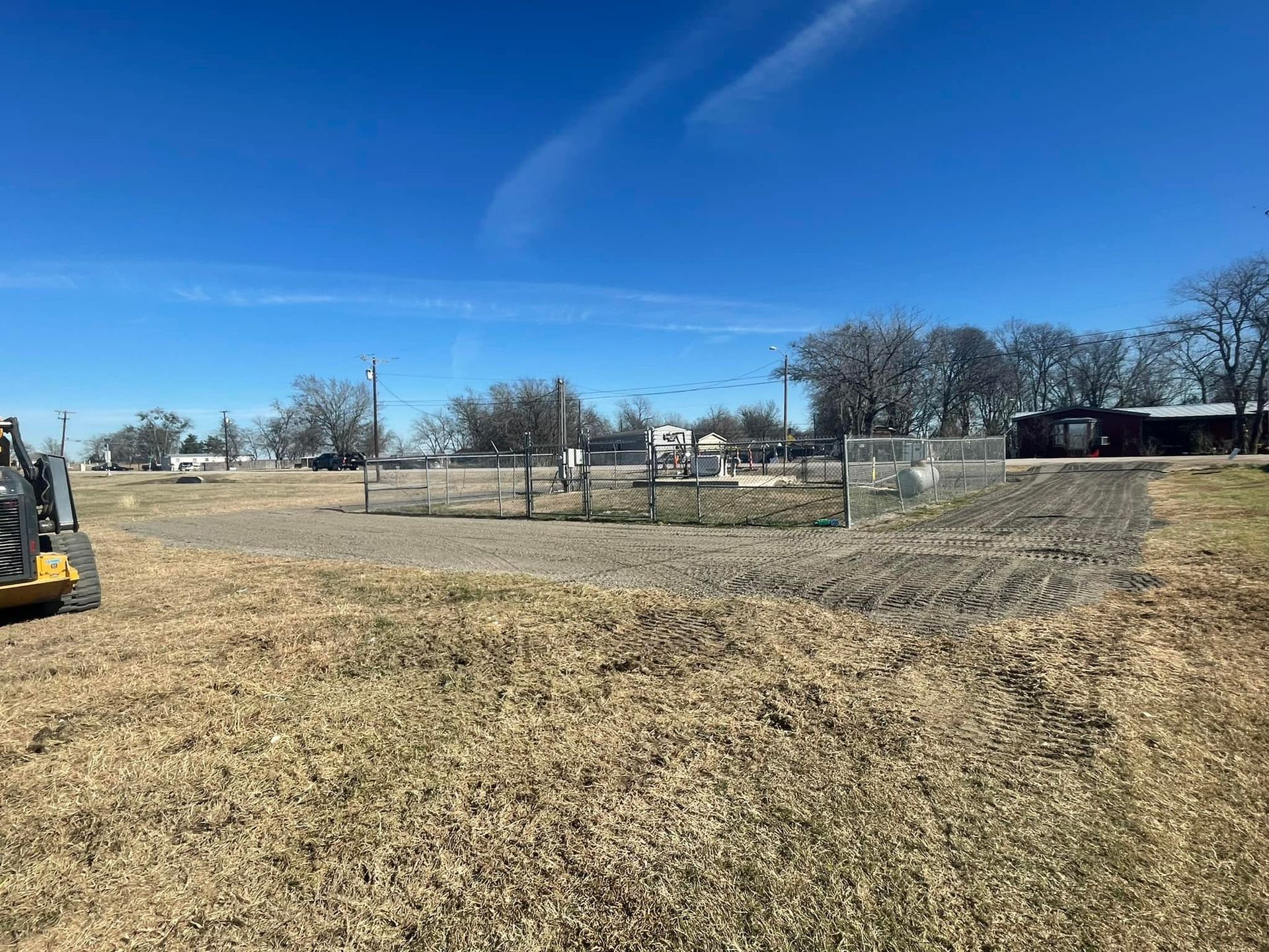 A field with brown grass and dirt, a metal fence, and a blue sky in a rural area.