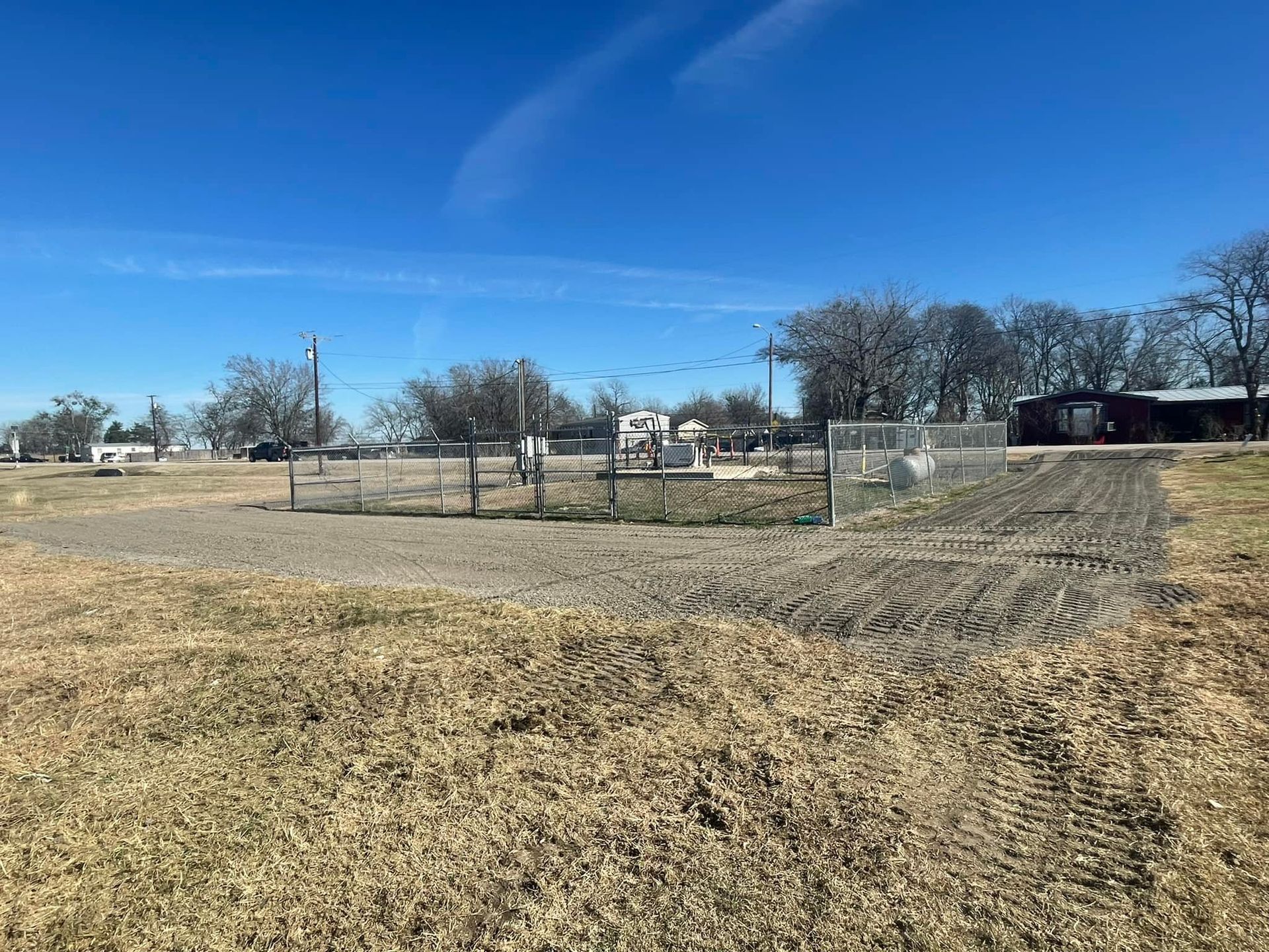 A fenced garden area on a dry field under a blue sky, small buildings and trees in background.