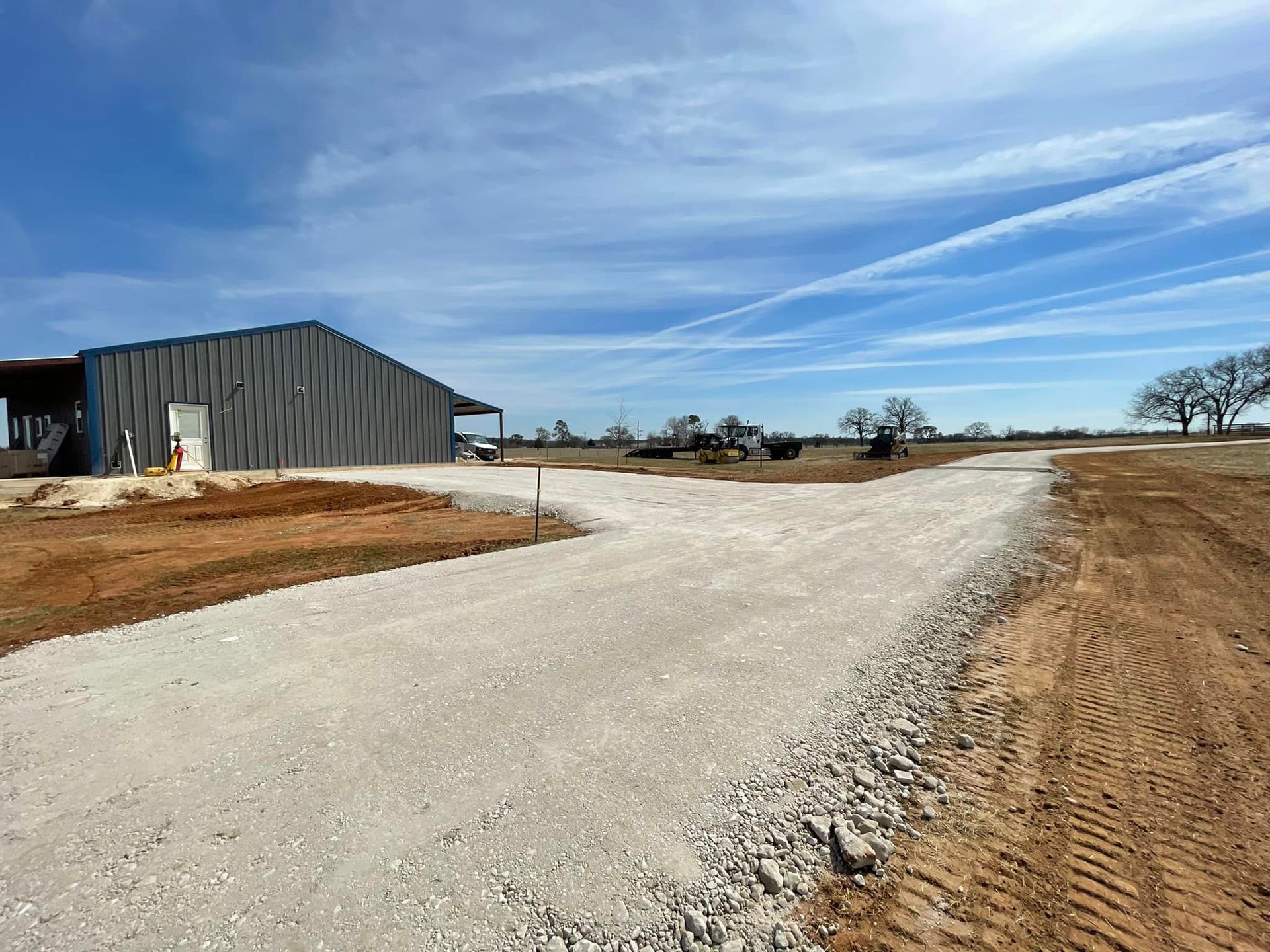 Gravel driveway leading to a metal building under a blue sky. Rural landscape with trees and dirt.