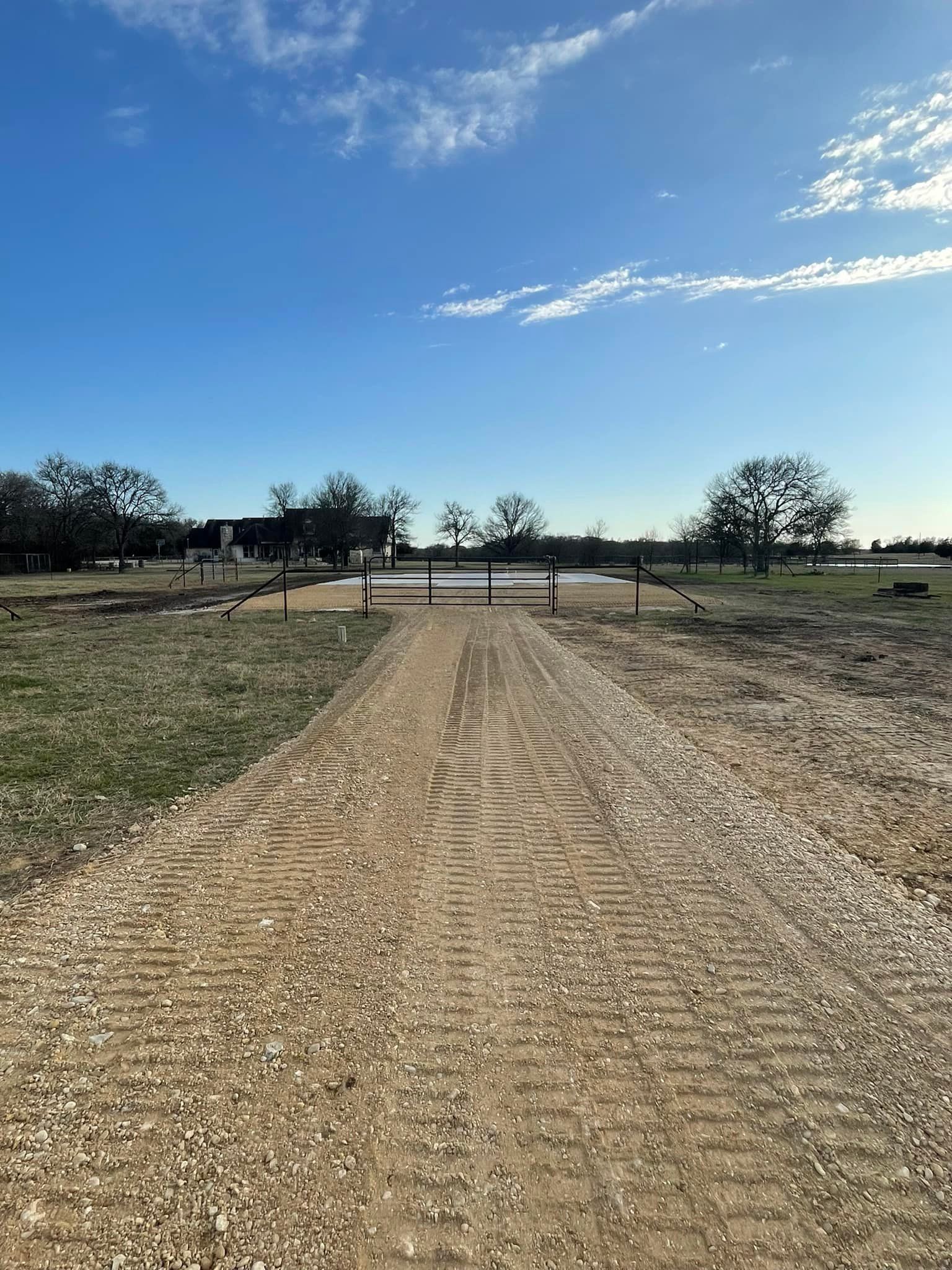 Dirt path leading toward a gate, open field, trees, and bright blue sky.