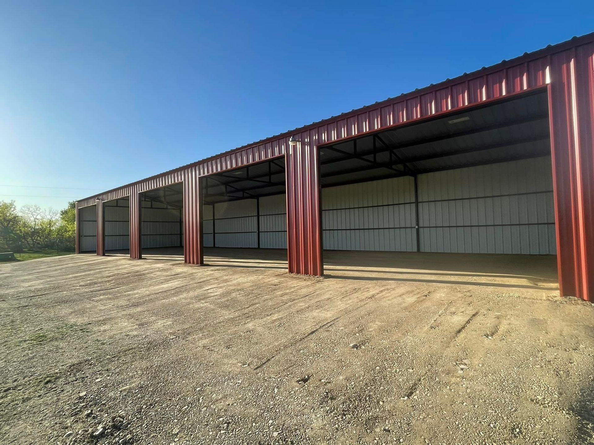 Red metal storage building with multiple open bays on a gravel surface under a blue sky.