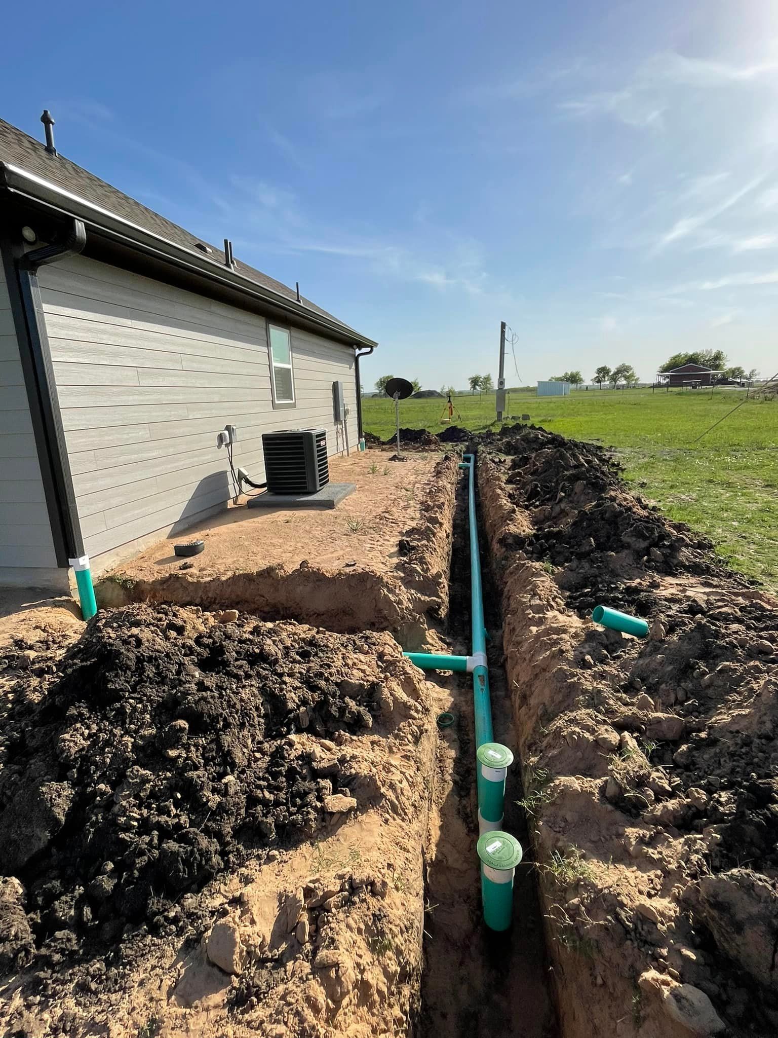 Trench with PVC pipes alongside a house; construction work outdoors on a sunny day.