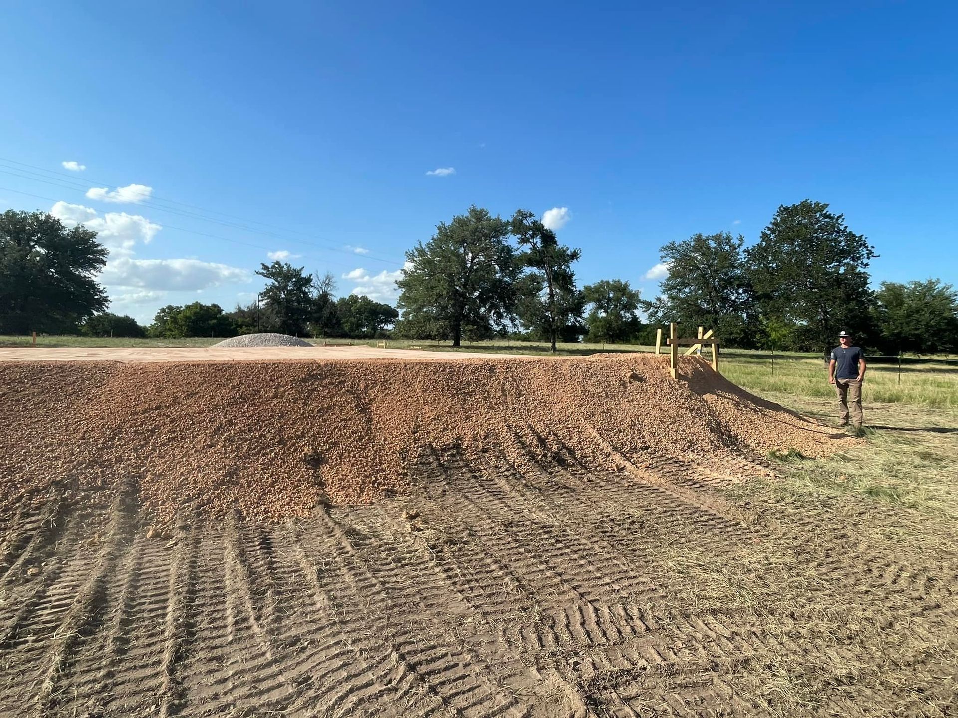 A dirt pile with a yellow ramp, a person walks nearby, trees and blue sky in the background.