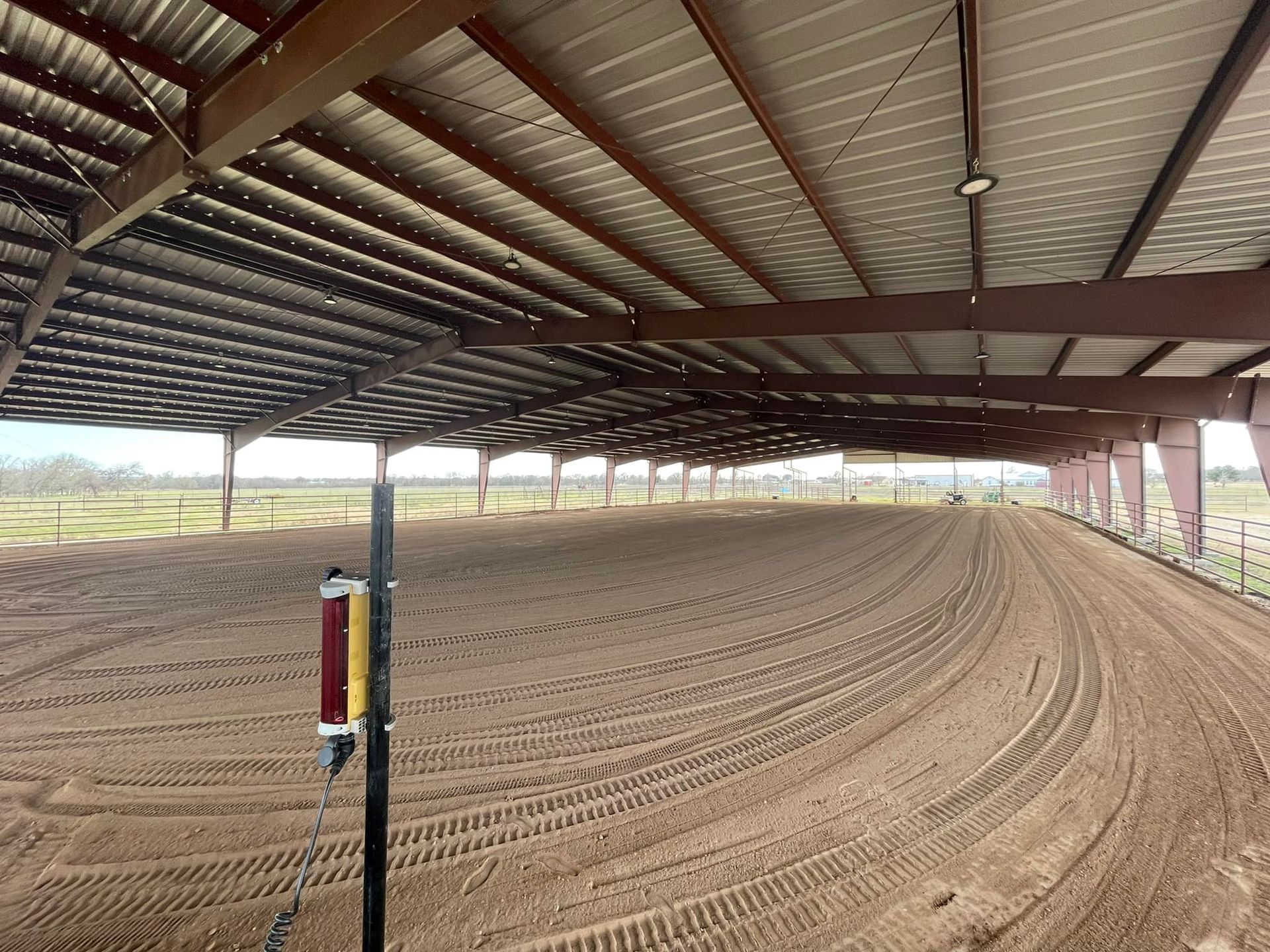 An open-air riding arena with brown dirt ground. Metal roof, brown supports, and a grassy field in the distance.
